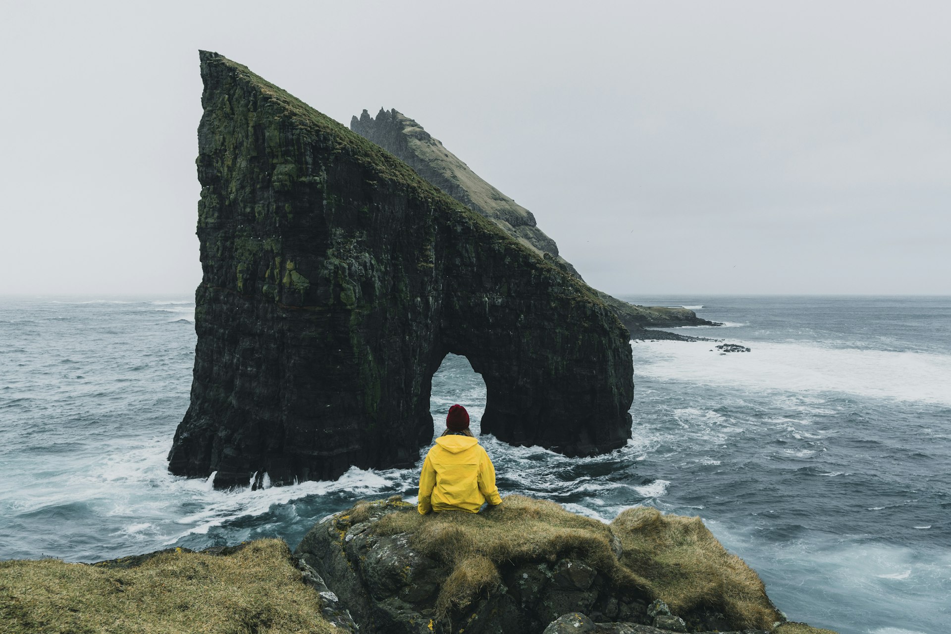 Woman in yellow raincoat looking at Drangarnir arch in Faroe Islands.