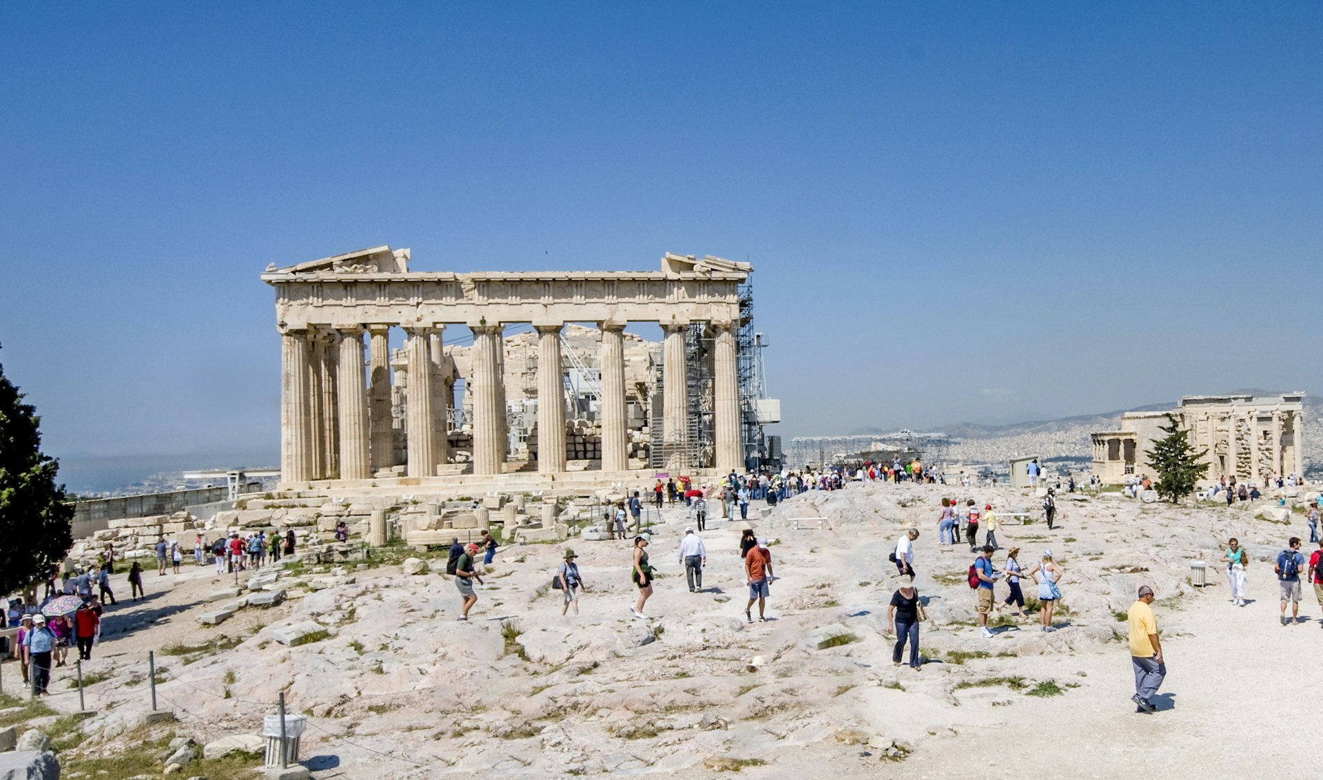 People follow paths through an archaeological site on a sunny day. The remains of a columned temple stand behind