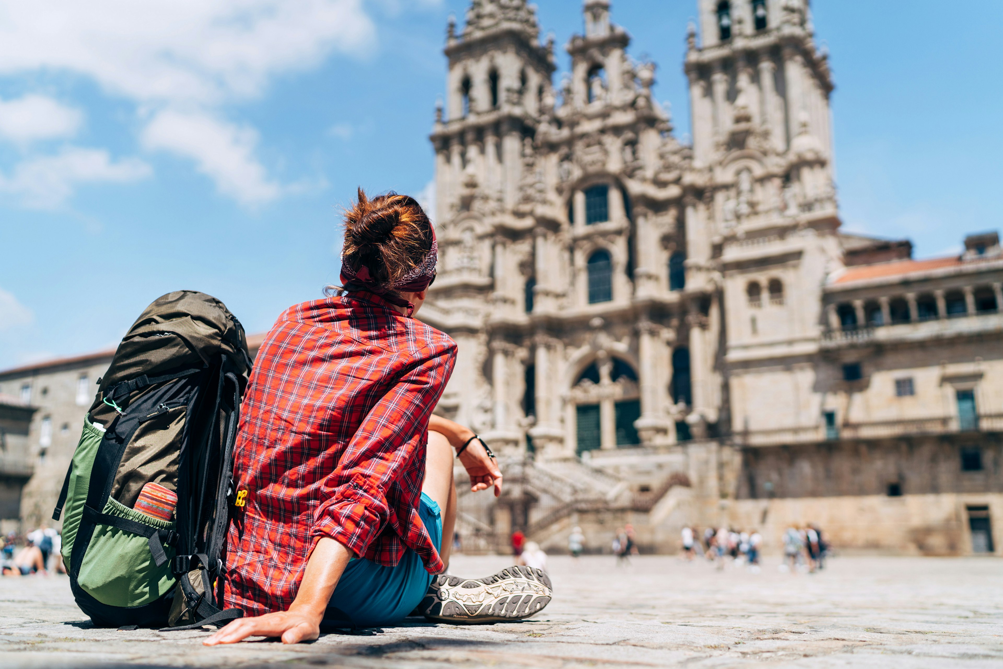 A pilgrim sits in a square with her backpack facing a large Gothic cathedral facade