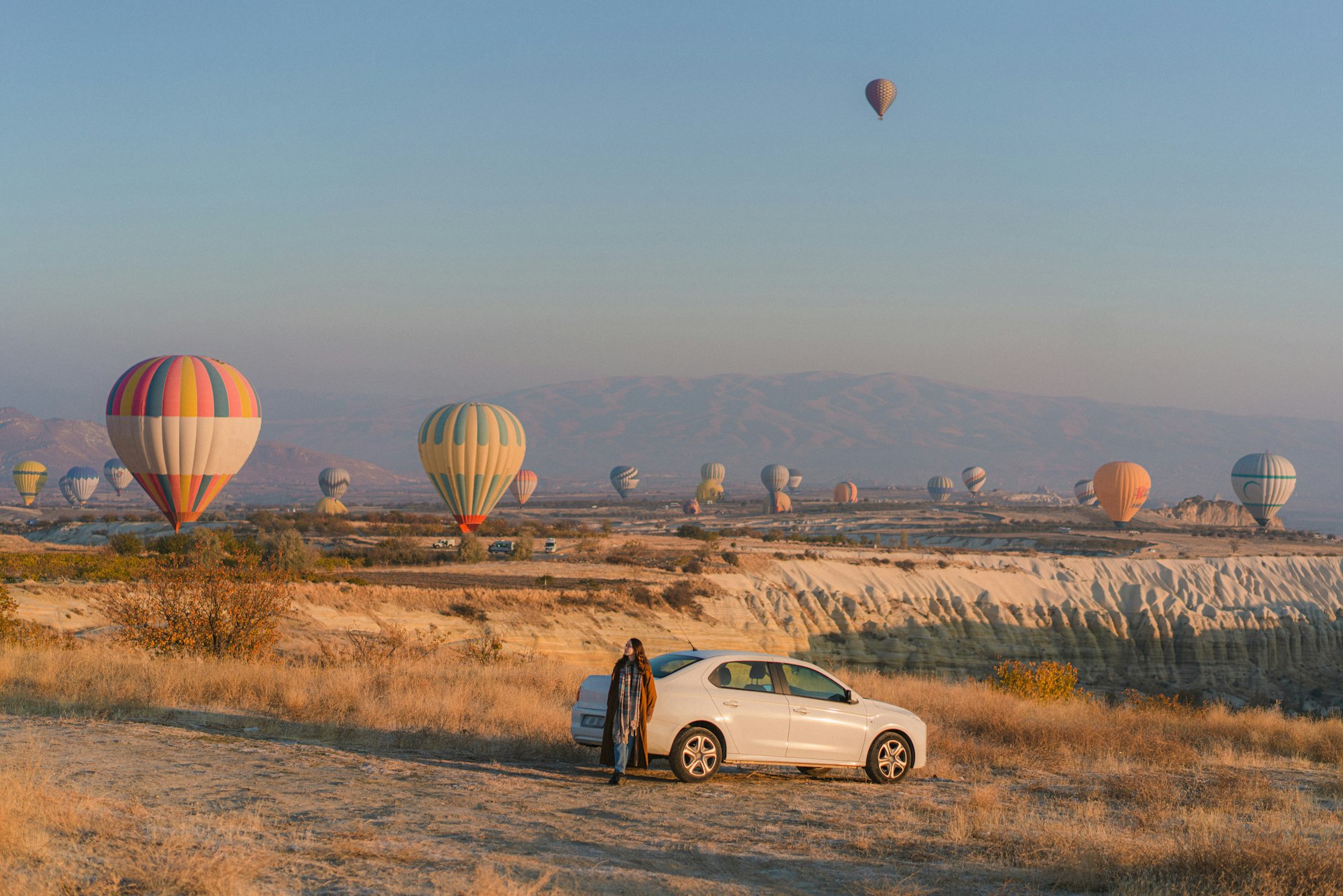 A woman sits against a white car parked in front of a field full of hot air balloons in Cappadocia