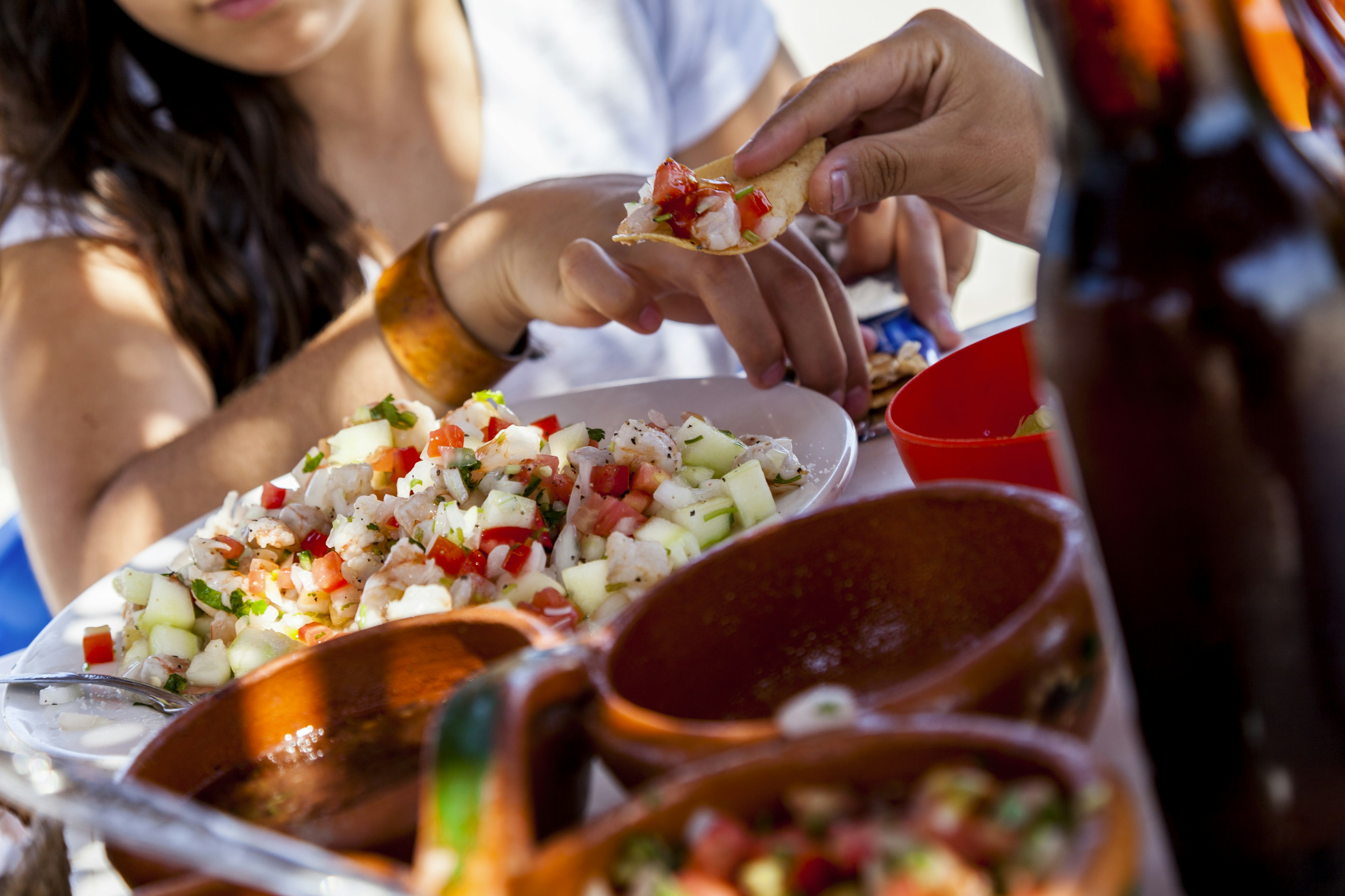 Two Hispanic Women friends sharing a Mexican lunch of seafood salad ceviche relaxing under a palapa on a beach in Ѳٱá, Mexico