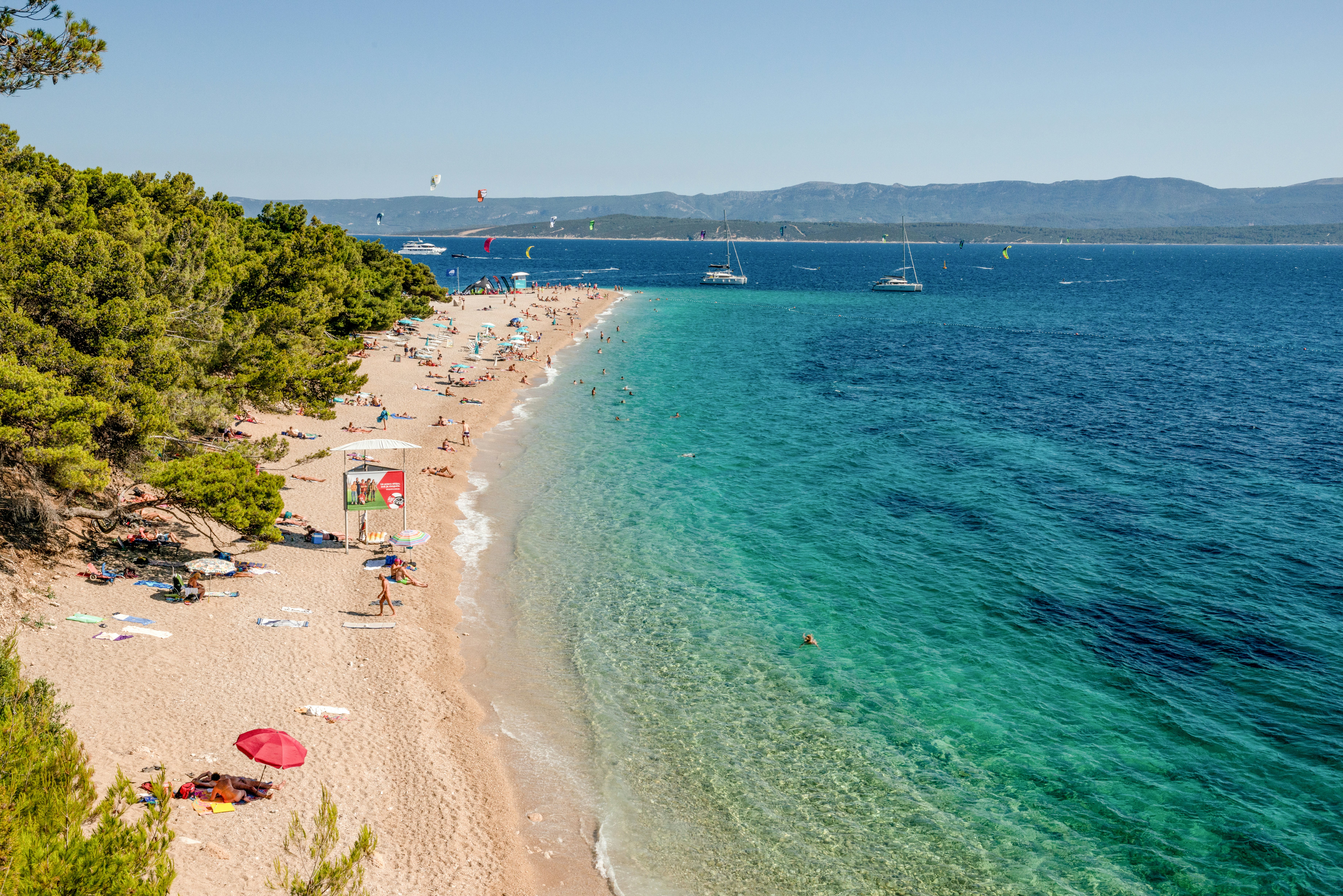 Tourists sunbathe on a beach backed by pine forest. Kitesurfers and yachts are in the distance.