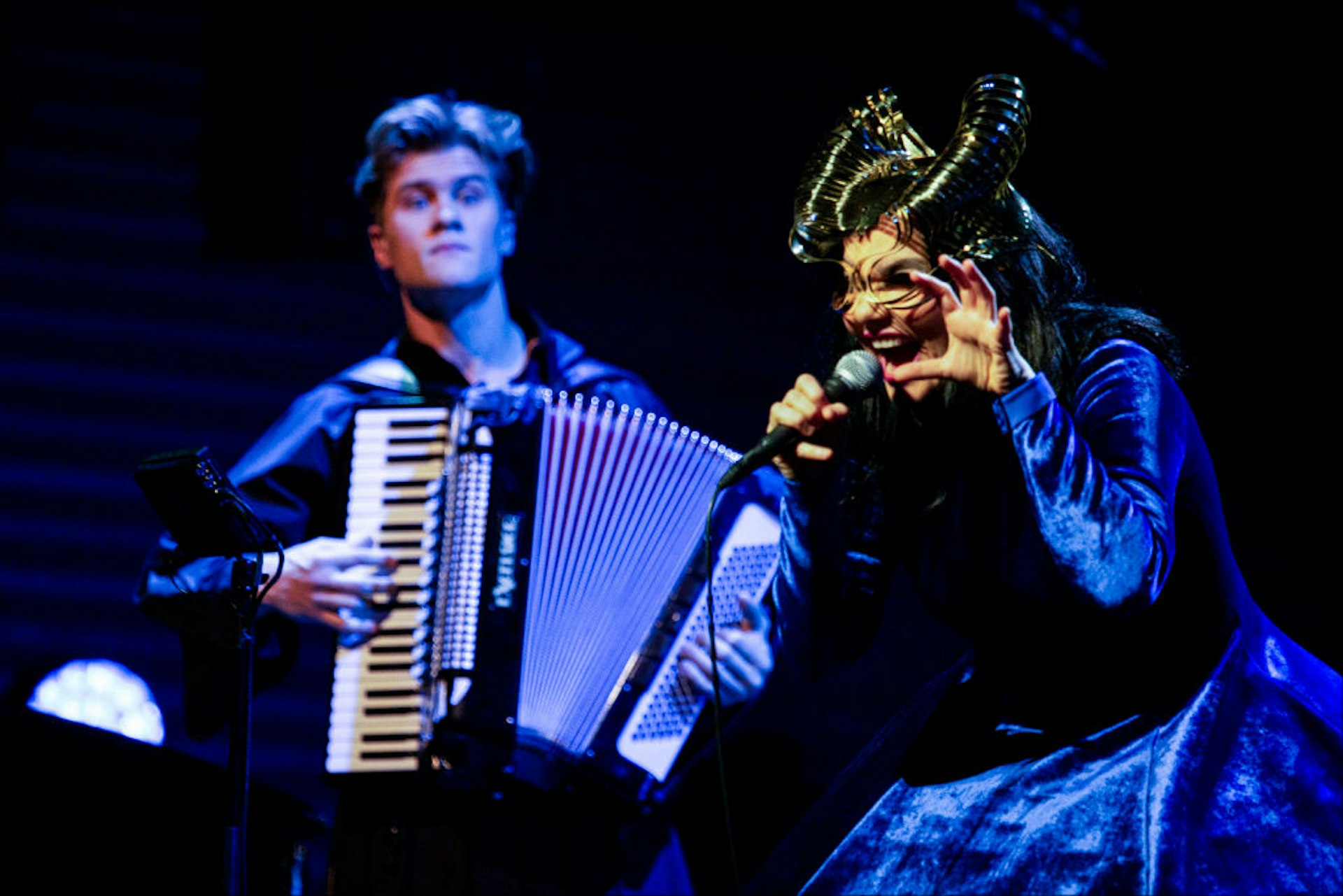 A woman wearing an elaborate face mask performs; an accordion player stands behind her