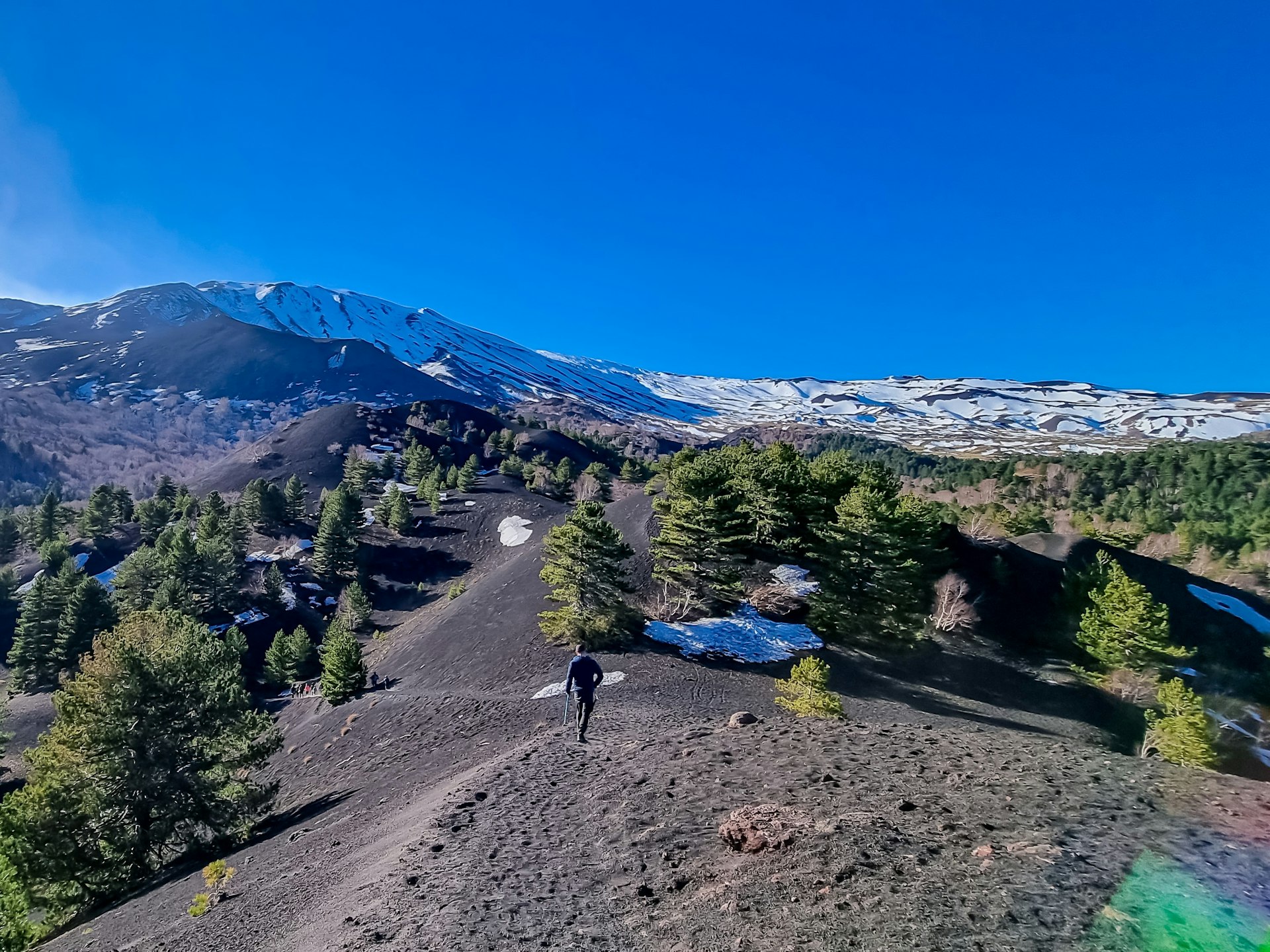 Um homem caminhando pela paisagem vulcânica do Monte Etna, na Sicília.  Pinheiros e bétulas brancas crescem sobre a lava solidificada, cinzas e pedra-pomes, e as encostas estão cobertas de neve.