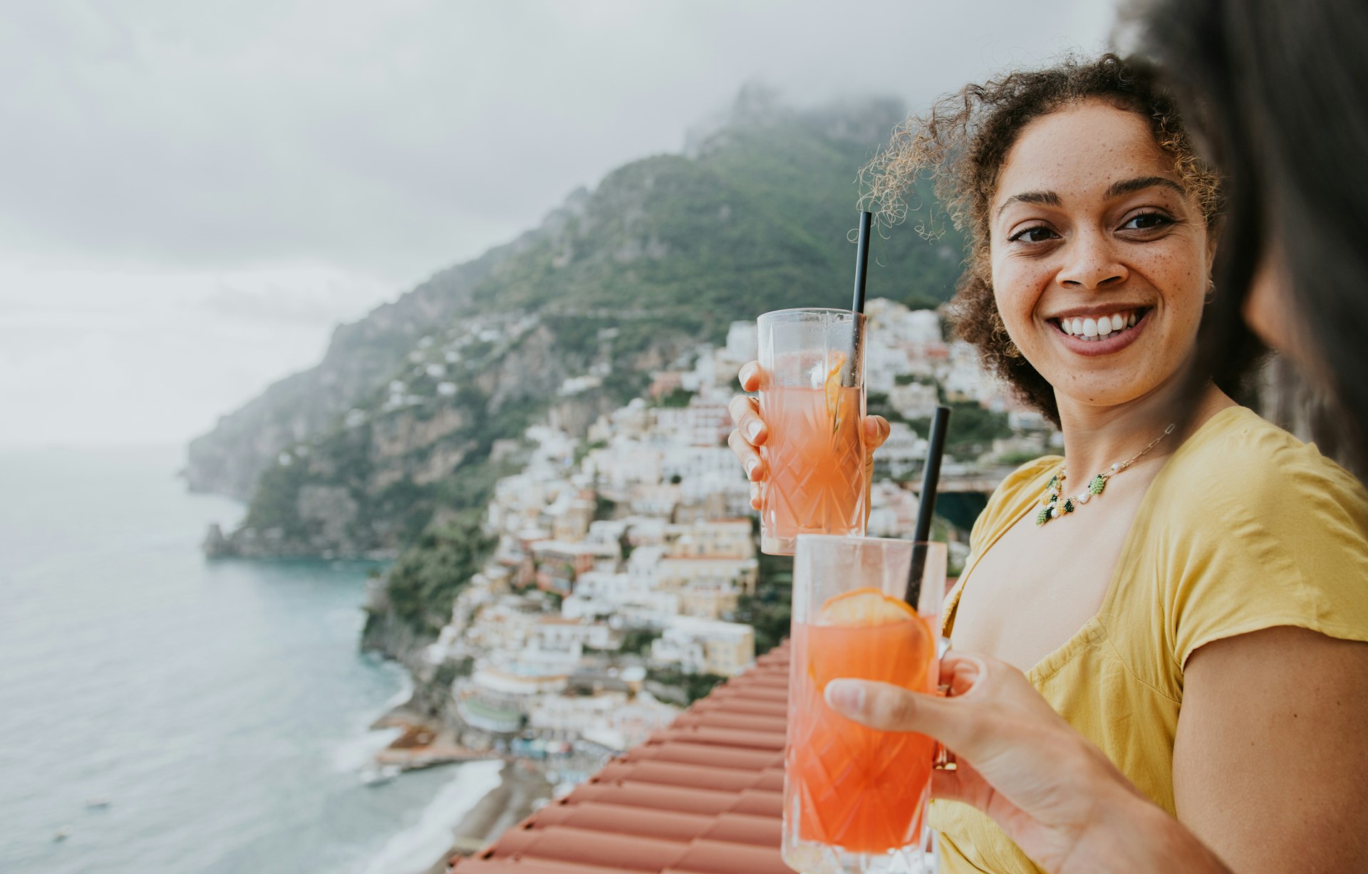 Two woman enjoy cocktails on a balcony overlooking Positano, Italy