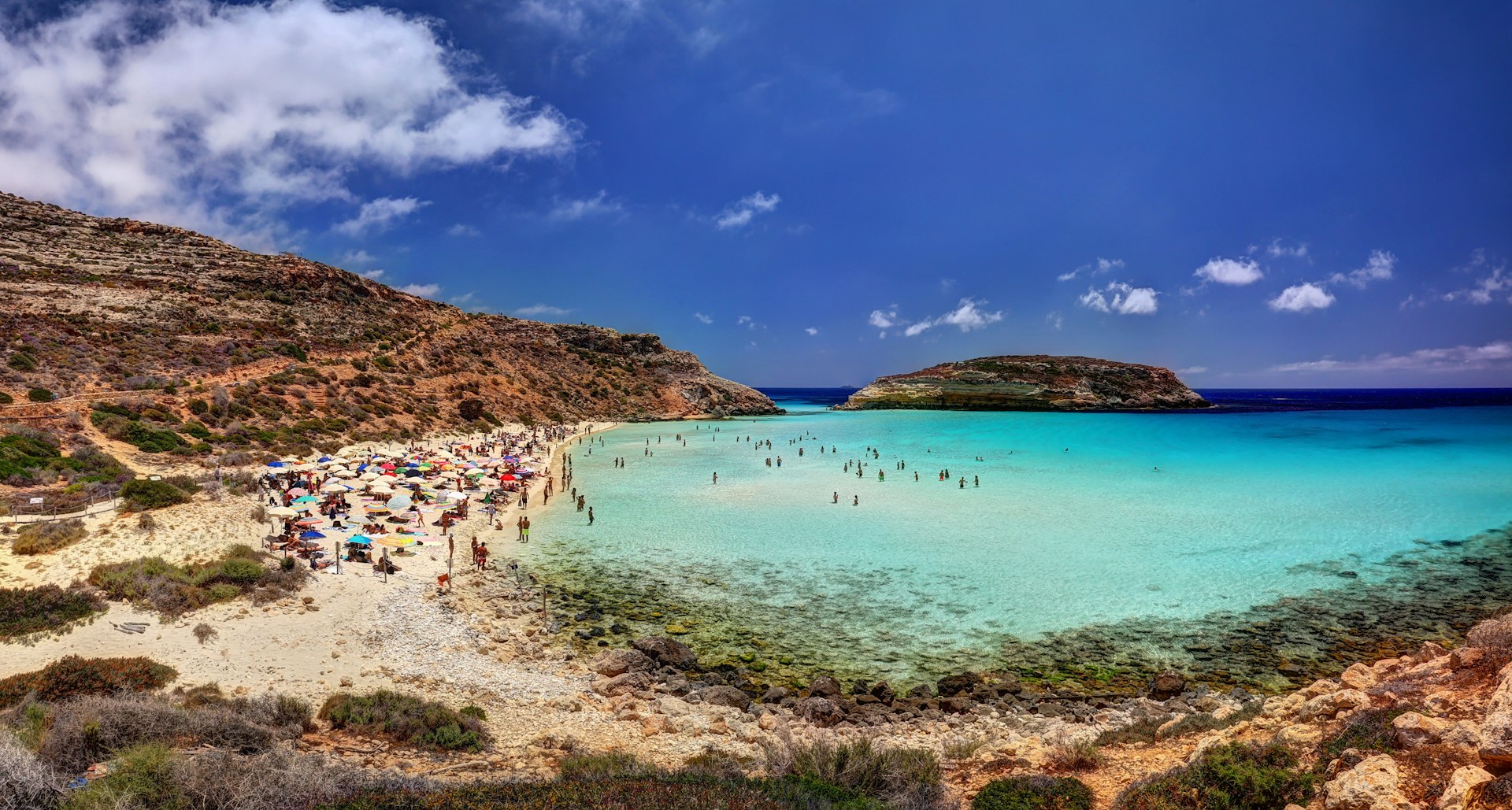 People swim in the turquoise cove at Rabbit Beach. The beach is surrounded by orange rock and there's a small island in the distance.