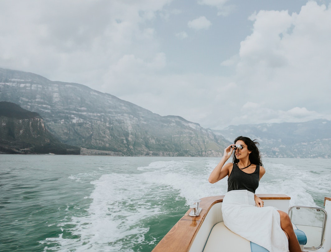 An elegant Indian woman sits on the back of a small, luxury yacht. As the boat moves along, she looks through a pair of silver binoculars. Admiring a view of mountains and sky.
1507780440
