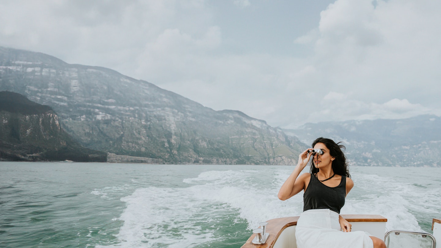 An elegant Indian woman sits on the back of a small, luxury yacht. As the boat moves along, she looks through a pair of silver binoculars. Admiring a view of mountains and sky.
1507780440