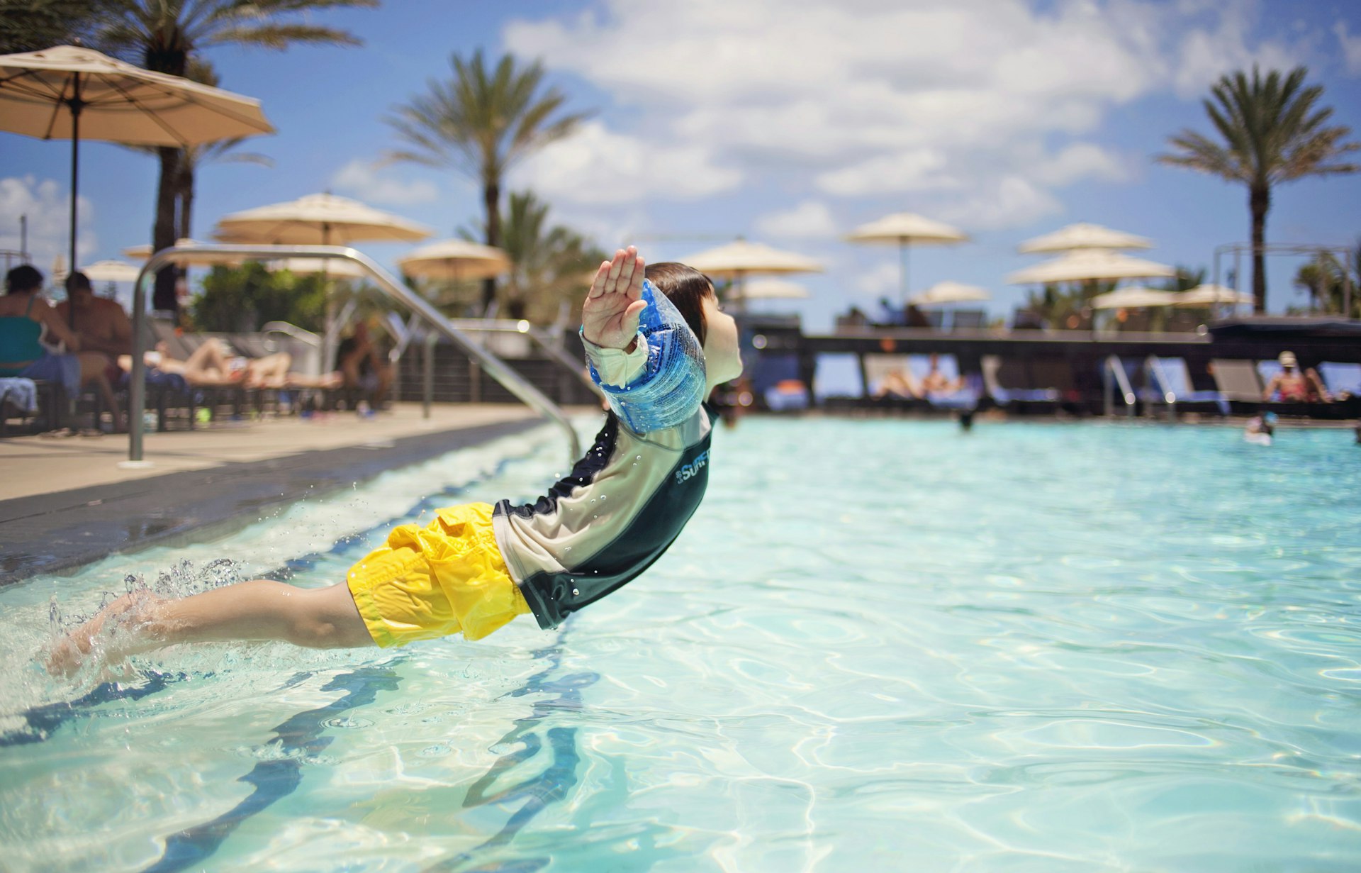 A small boy wearing water wings jumps into a hotel pool that's surrounded by sun loungers