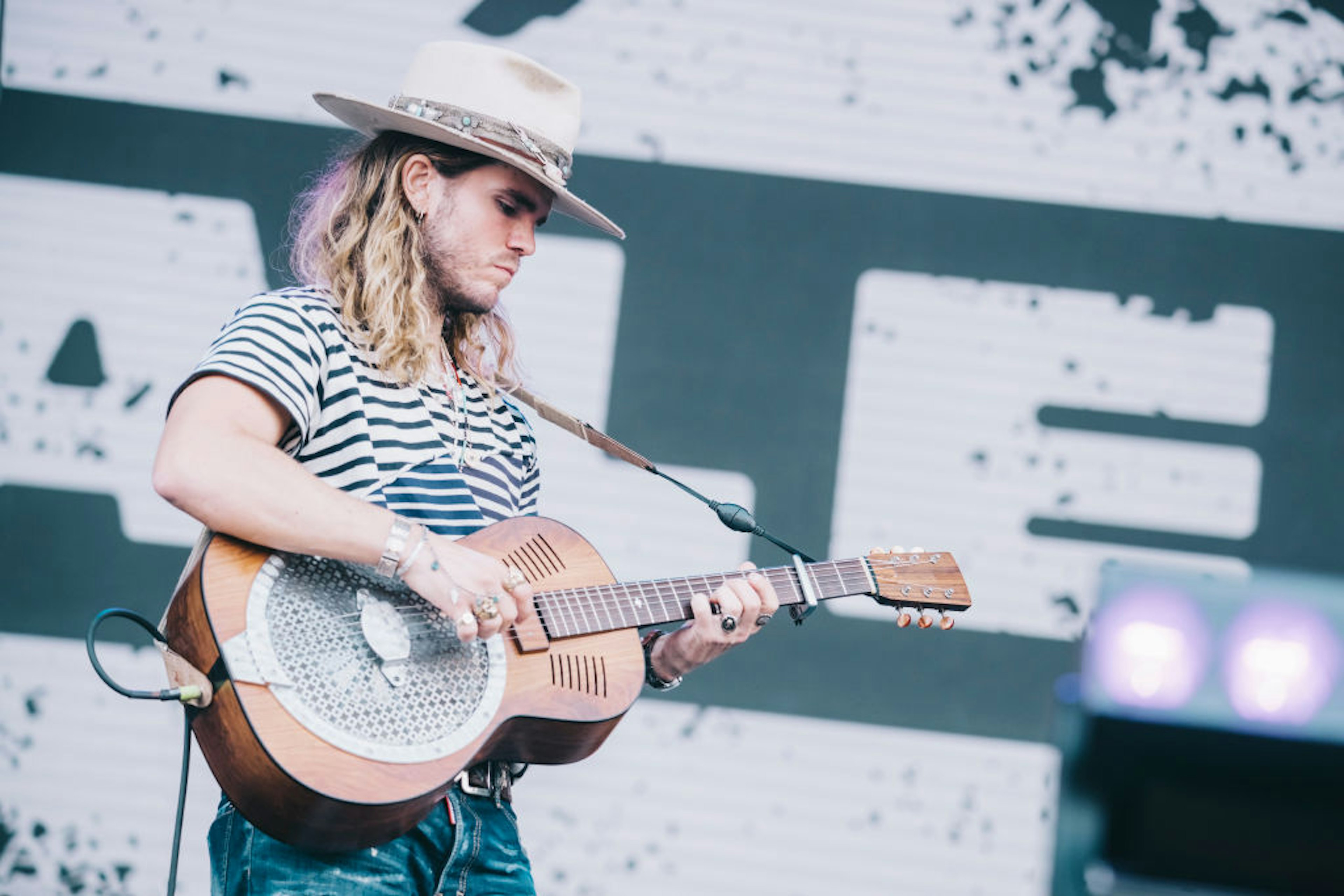 A guitarist with long hair and a cowboy-style hat plays guitar on stage