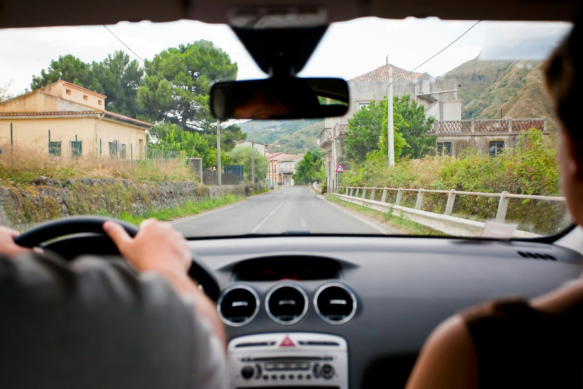 A man and woman sit in a car while driving in Sicily. The camera is looking out the front windshield to reveal a historic town ahead