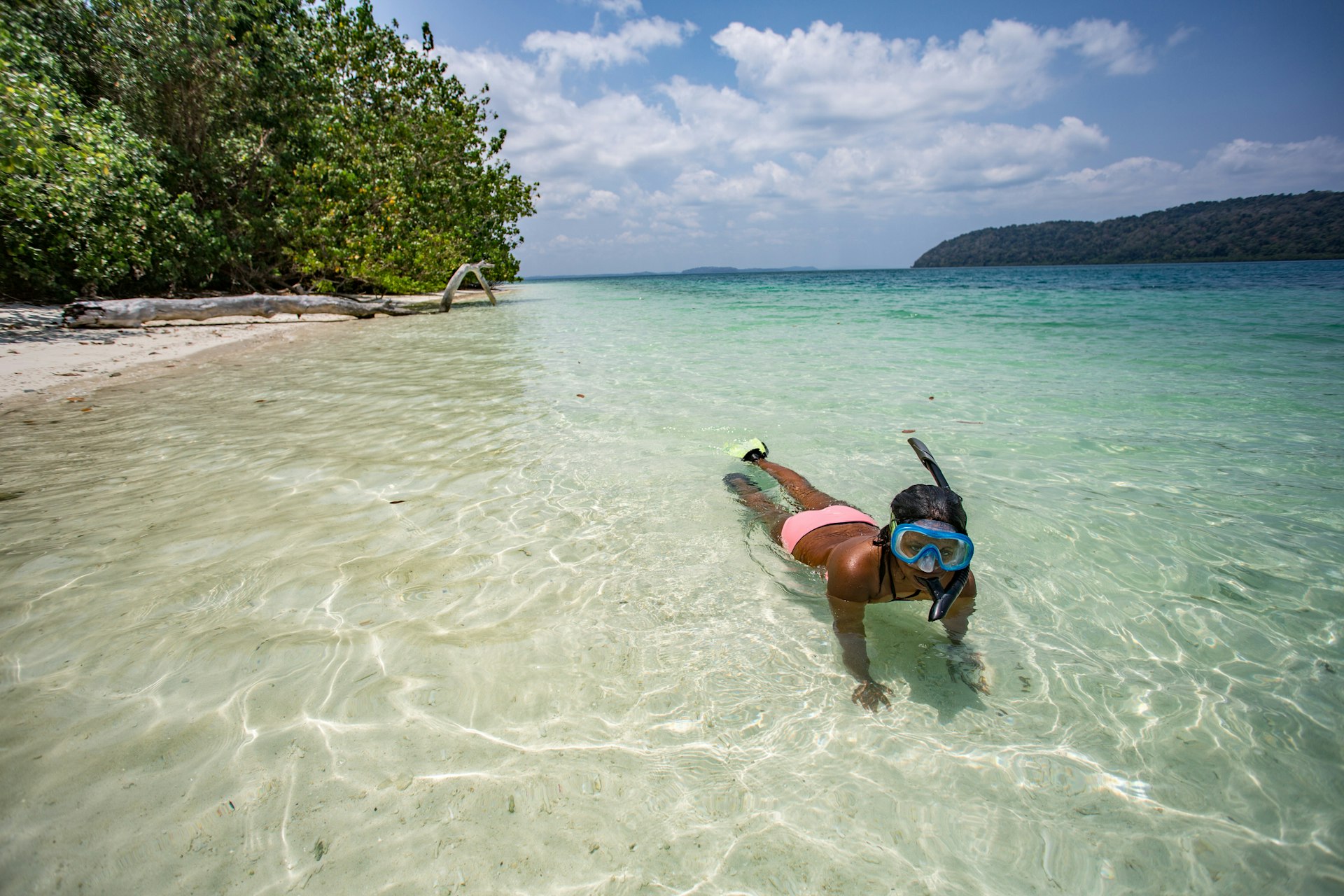A girl with mask and snorkel swims in shallow clear waters just off a beach