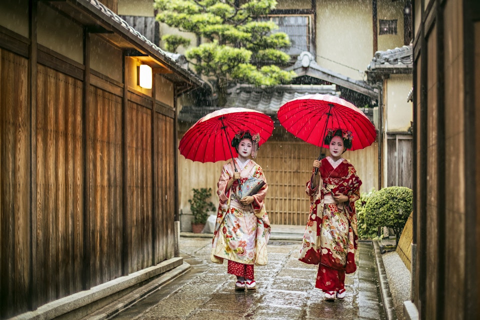 Full length of young maikos holding red umbrellas during rainy season. Beautiful geisha girls wearing traditional dress called kimono. They are walking on wet street.
641625768