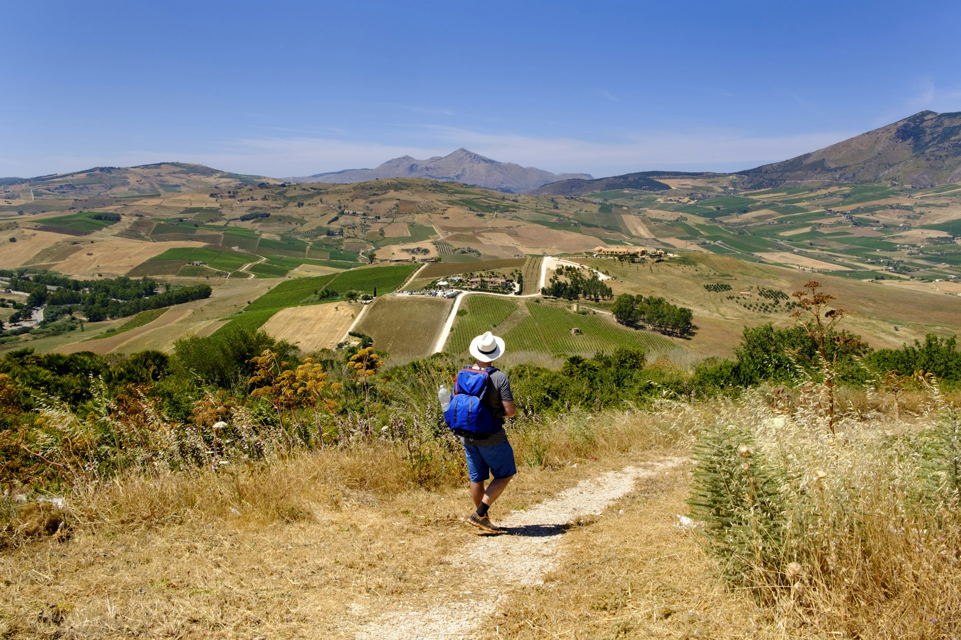 Uma pessoa usando um chapéu branco caminha por uma trilha com vista para a zona rural perto de Segesta, Itália