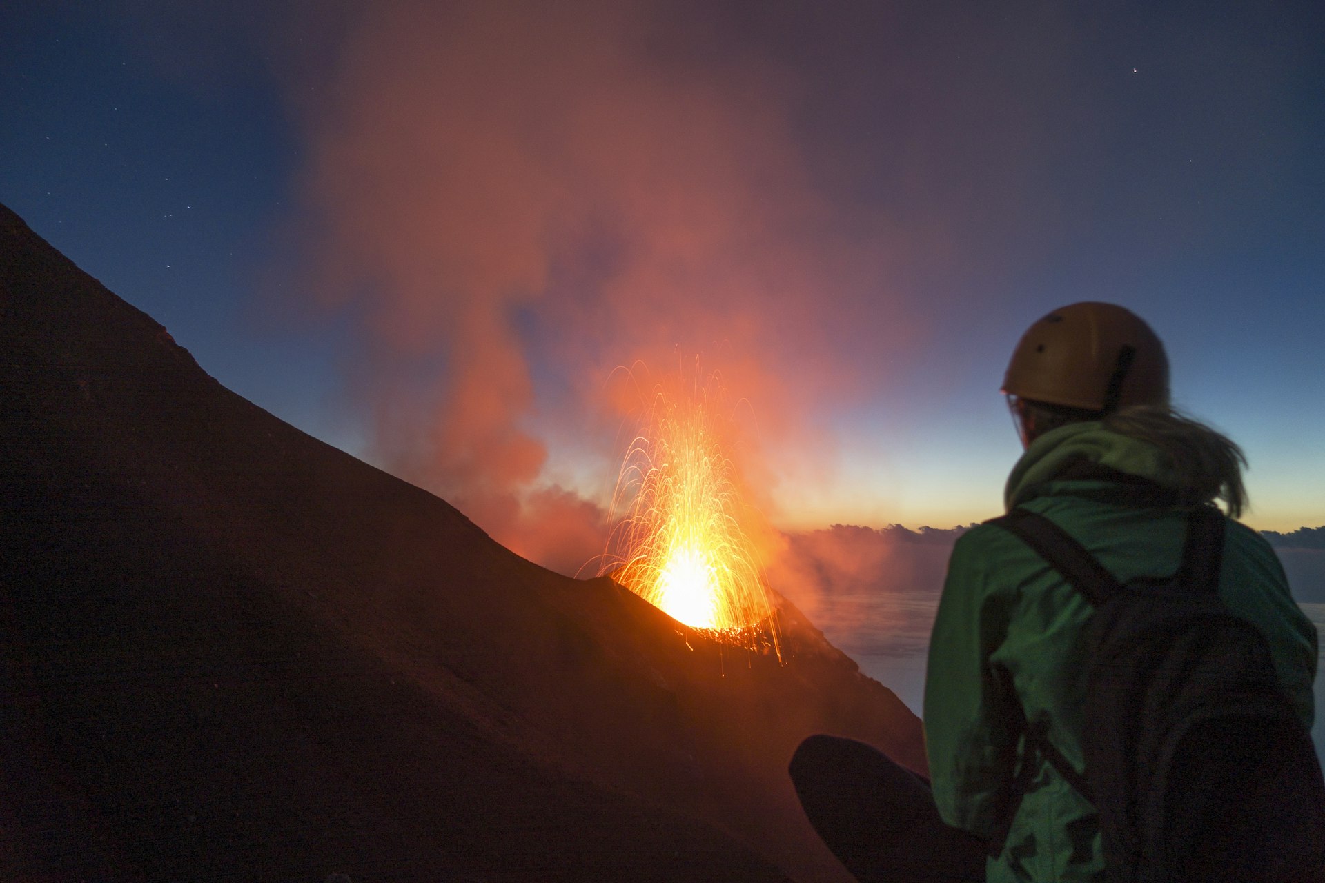 A woman in a helmet looks out over an erupting volcano at sunrise