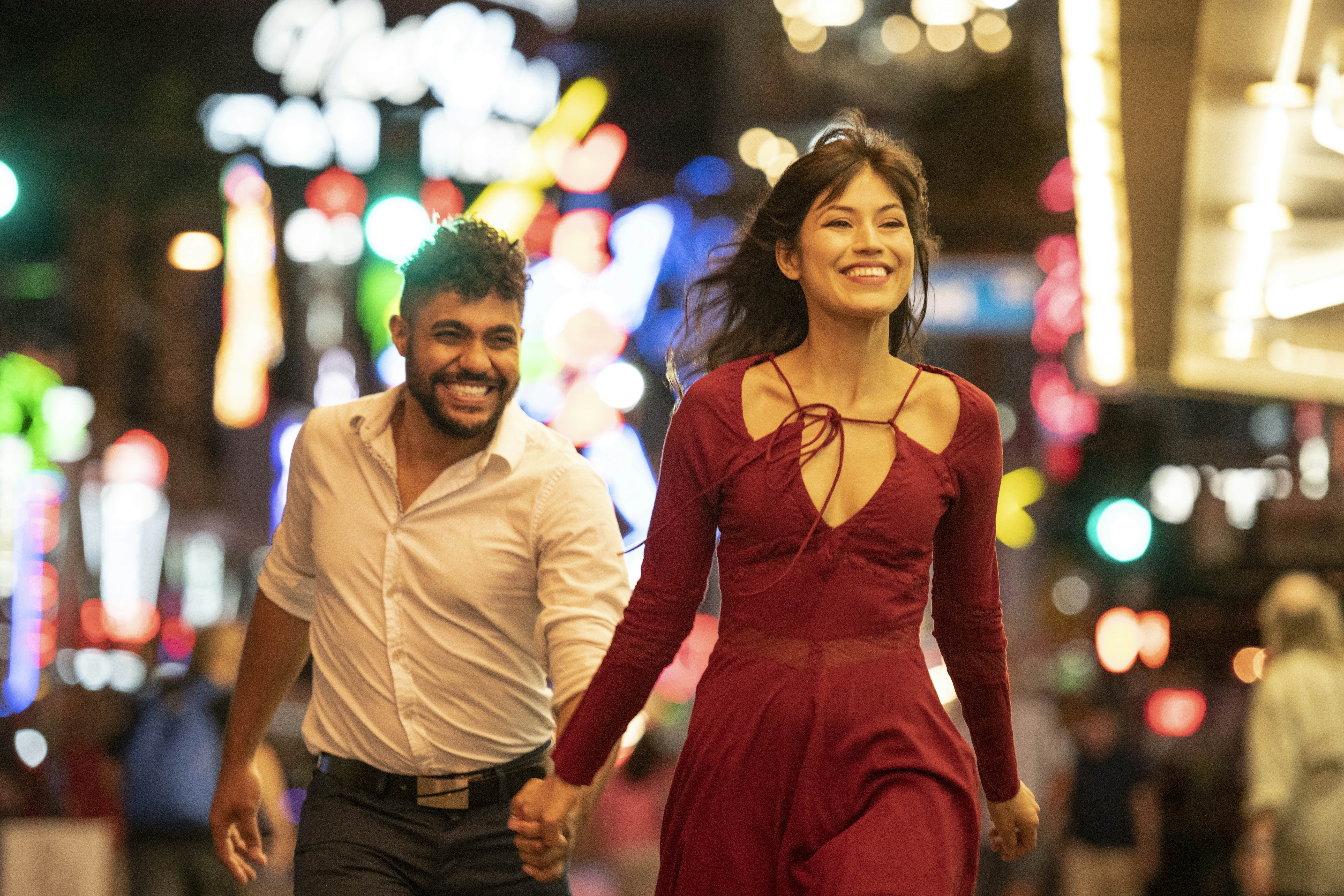 Latin American woman and mixed race man, in their 20's, wearing party clothes, having fun together with woman walking ahead smiling and bright city lights in background
994425004
