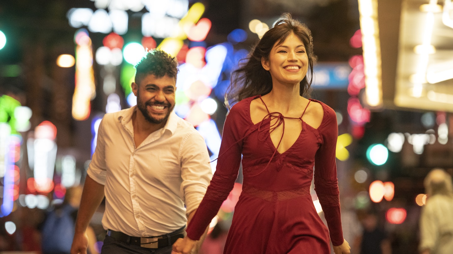 Latin American woman and mixed race man, in their 20's, wearing party clothes, having fun together with woman walking ahead smiling and bright city lights in background
994425004