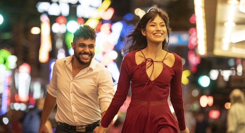 Latin American woman and mixed race man, in their 20's, wearing party clothes, having fun together with woman walking ahead smiling and bright city lights in background
994425004