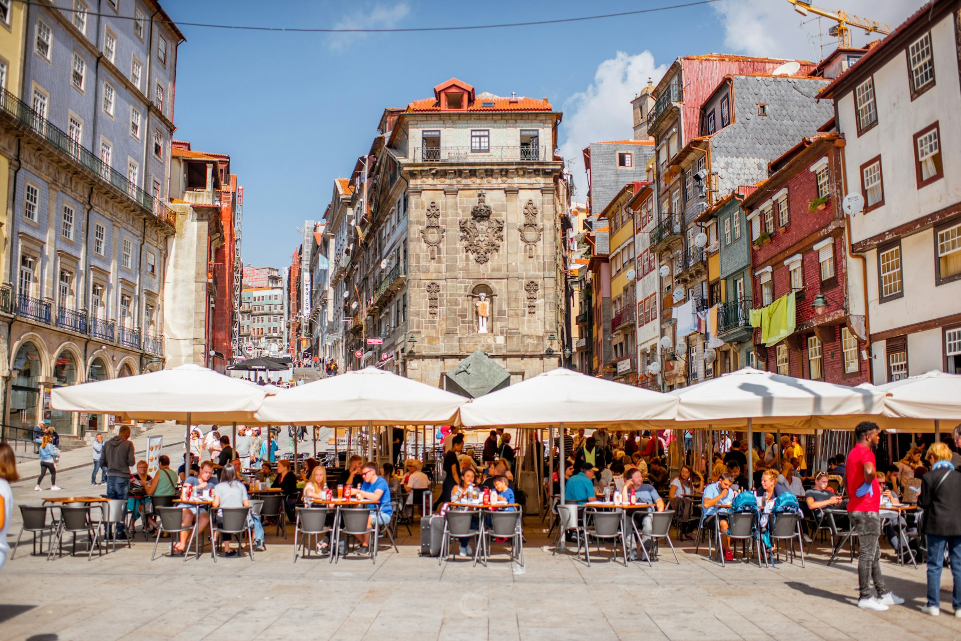 People sat at restaurant tables under sunshades in a city square