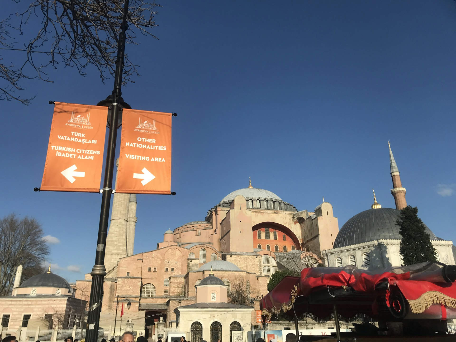  Image of a signpost outside Hagia Sophia with two orange signs. The left sign points to the left, indicating the entrance for "Turkish Citizens İbadet Alanı," while the right sign points to the right for "Other Nationalities Visiting Area." The background shows the exterior of Hagia Sophia with its distinctive domes and minarets against a clear blue sky.