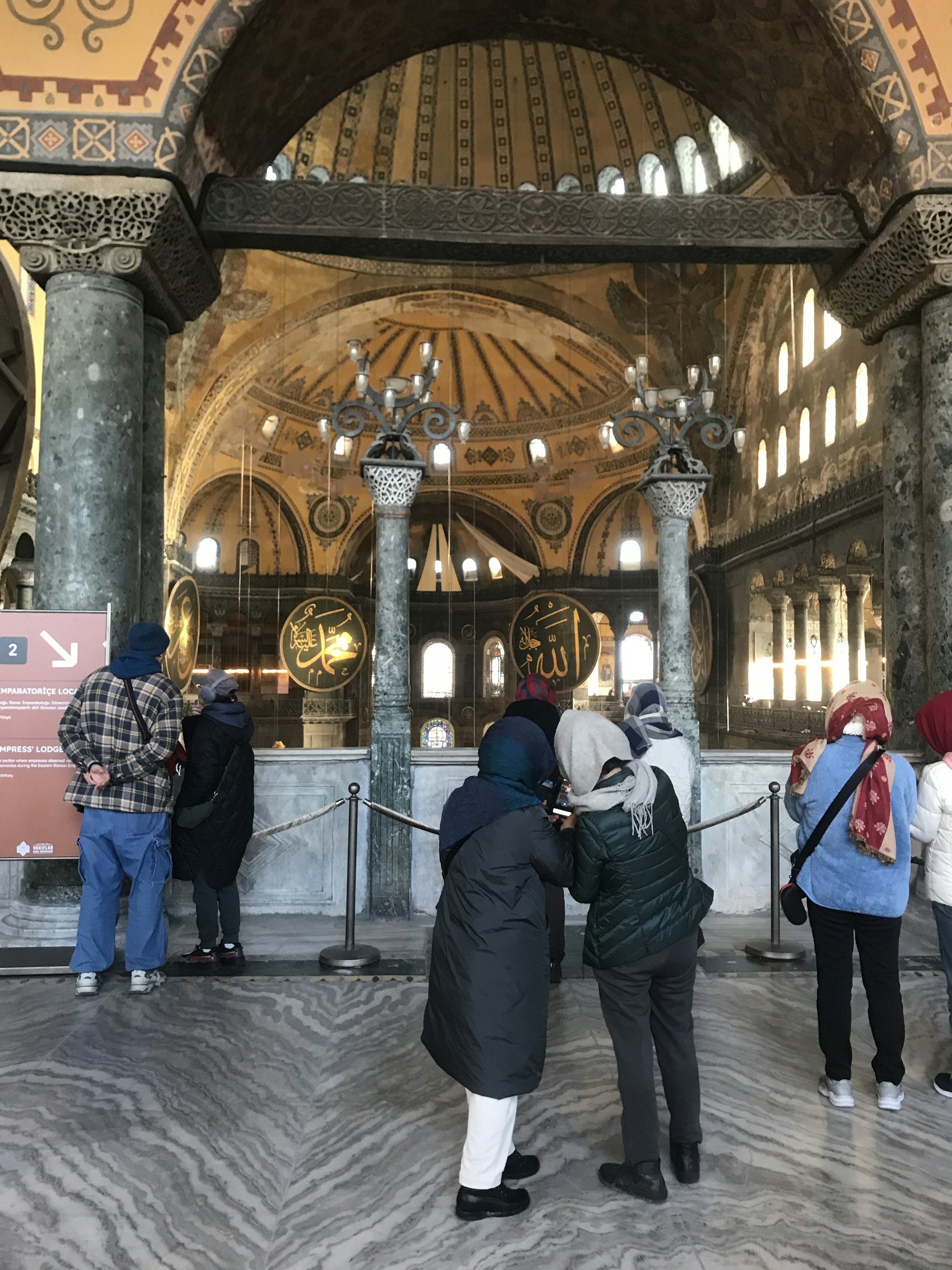  Image of the interior of Hagia Sophia where visitors are viewing the historical site. Several columns with decorative capitals and an ornate upper gallery with Islamic calligraphy are visible. Visitors are using audio guides, and the floor features a marble pattern. There's a roped-off section leading to an upper area of the mosque..jpg