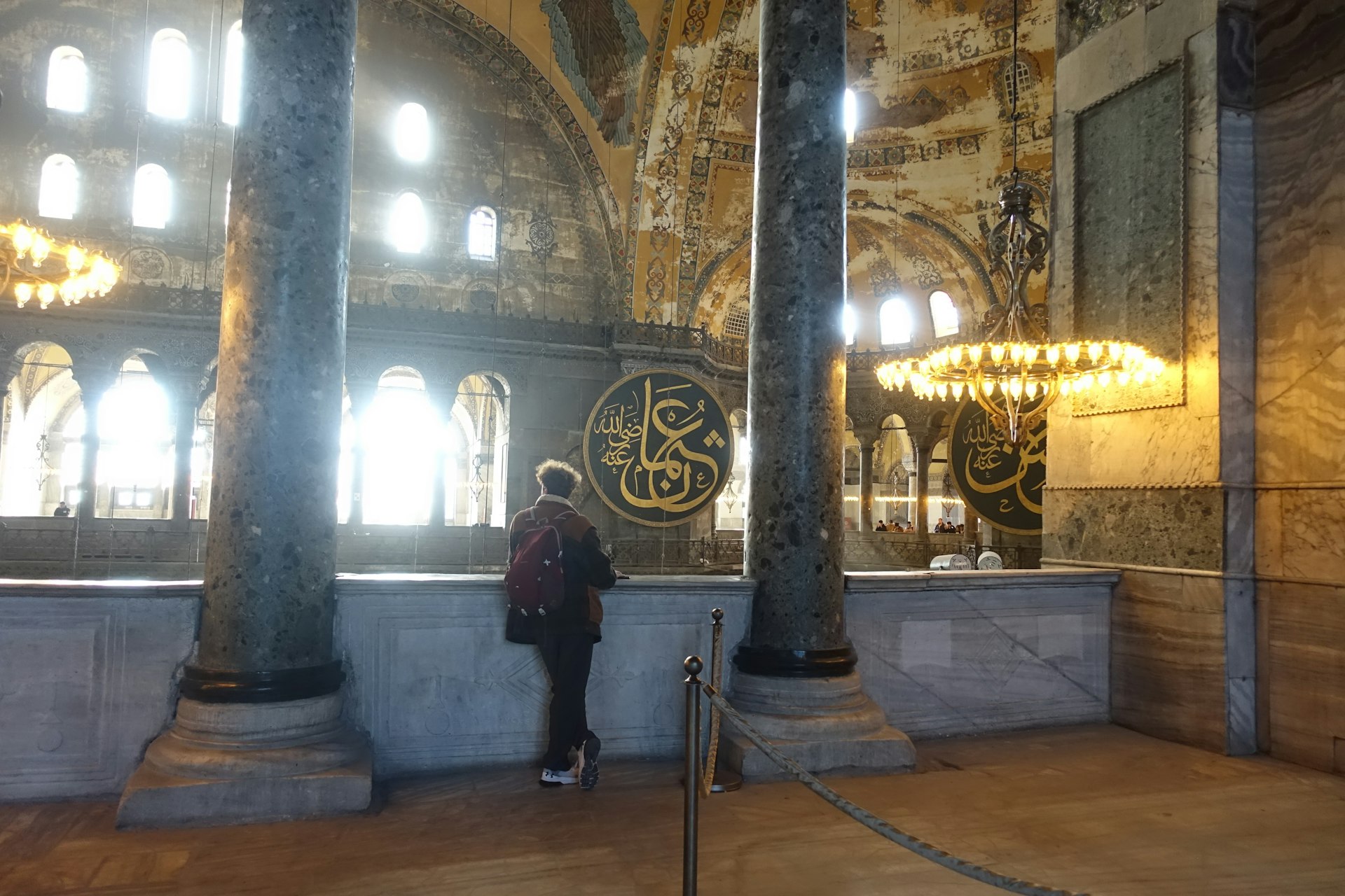 Image of the interior of Hagia Sophia showing a visitor observing the space. Large columns and a circular chandelier are visible, with Islamic calligraphic panels mounted on the walls. The ceilings showcase intricate designs, and the area is illuminated by natural light from the windows.