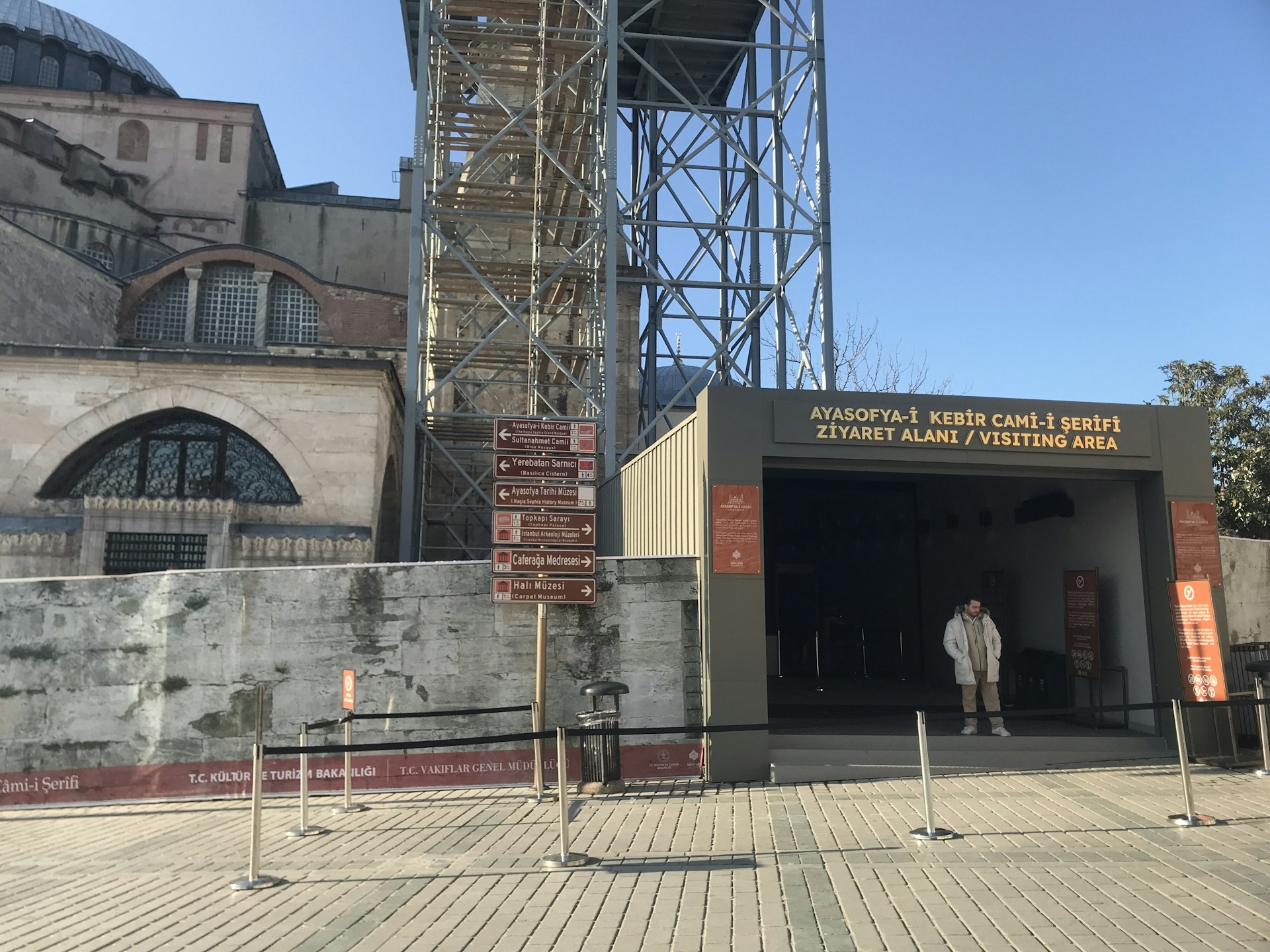Image of an entrance labeled "AYASOFYA-İ KEBİR CAMİ-İ ŞERİFİ ZİYARET ALANI / VISITING AREA." The entrance is a simple, modern design with a man standing in the doorway and scaffolding visible to the left, indicating possible restoration work. The sign above the entrance is in both Turkish and English..jpg