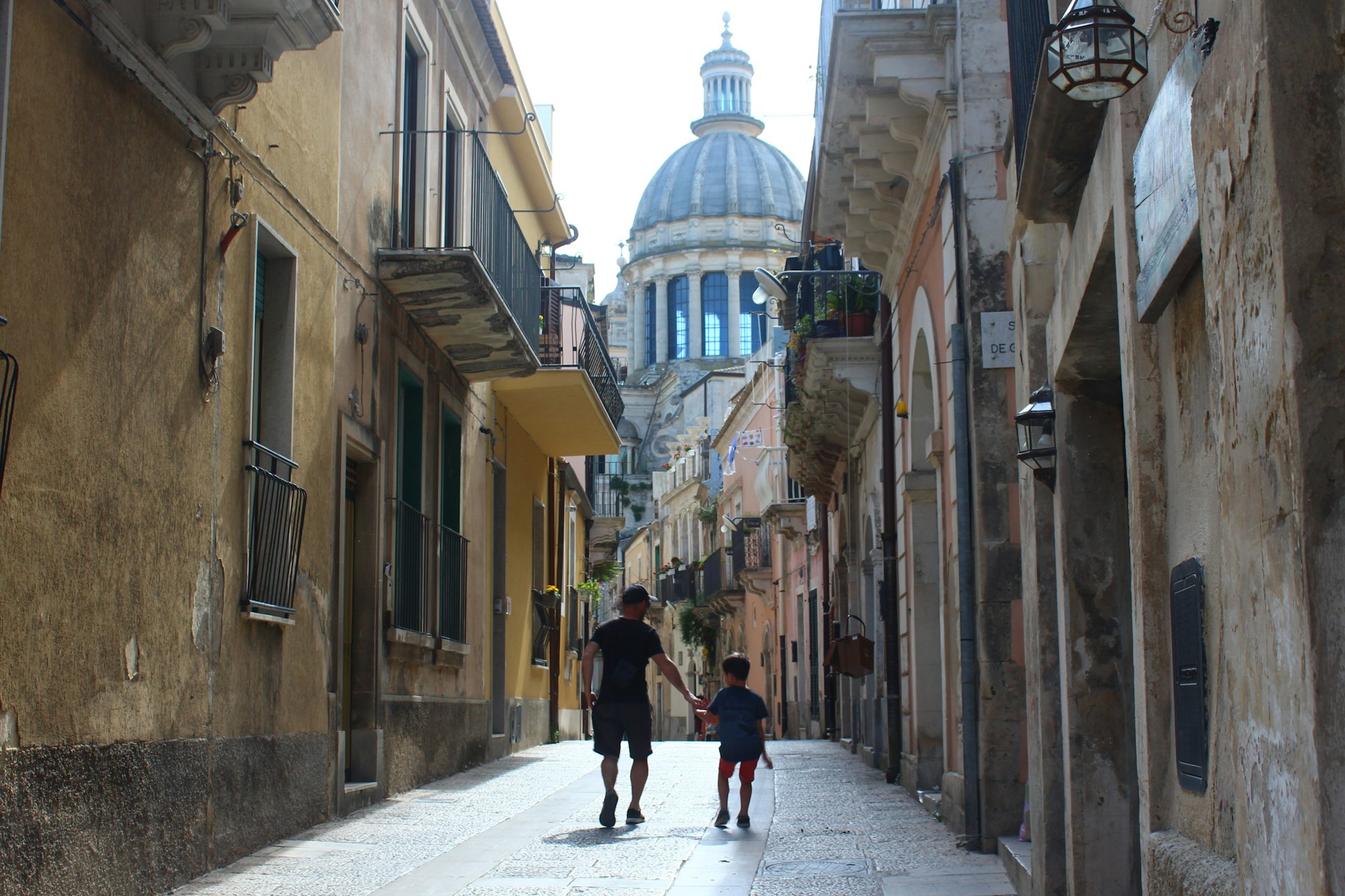 Pai e filho caminham por uma viela histórica em Ragusa, na Sicília, com a cúpula do Duomo di San Giorgio visível sobre os telhados
