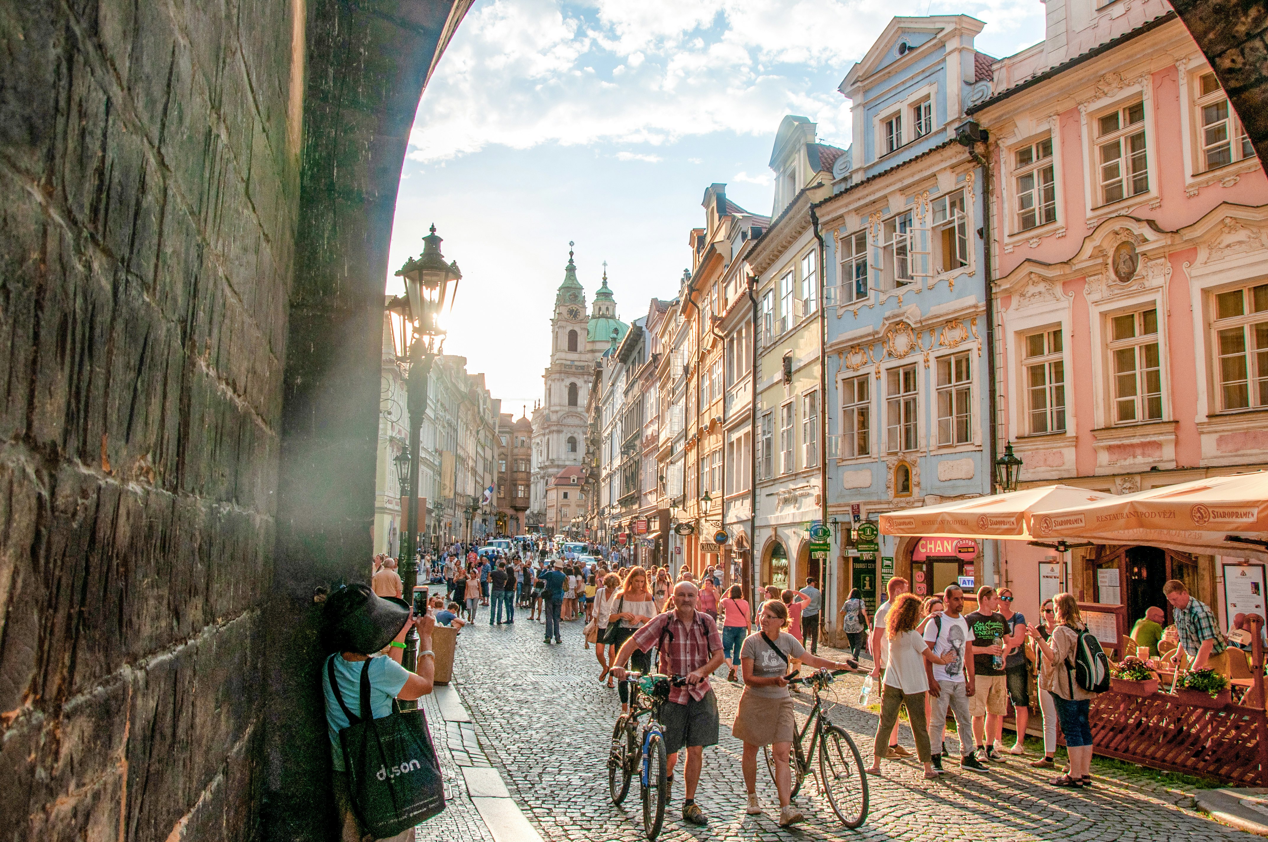 A crowded cobblestone street at sunset in the summer with lots of buildings & cyclists in the city of Prague