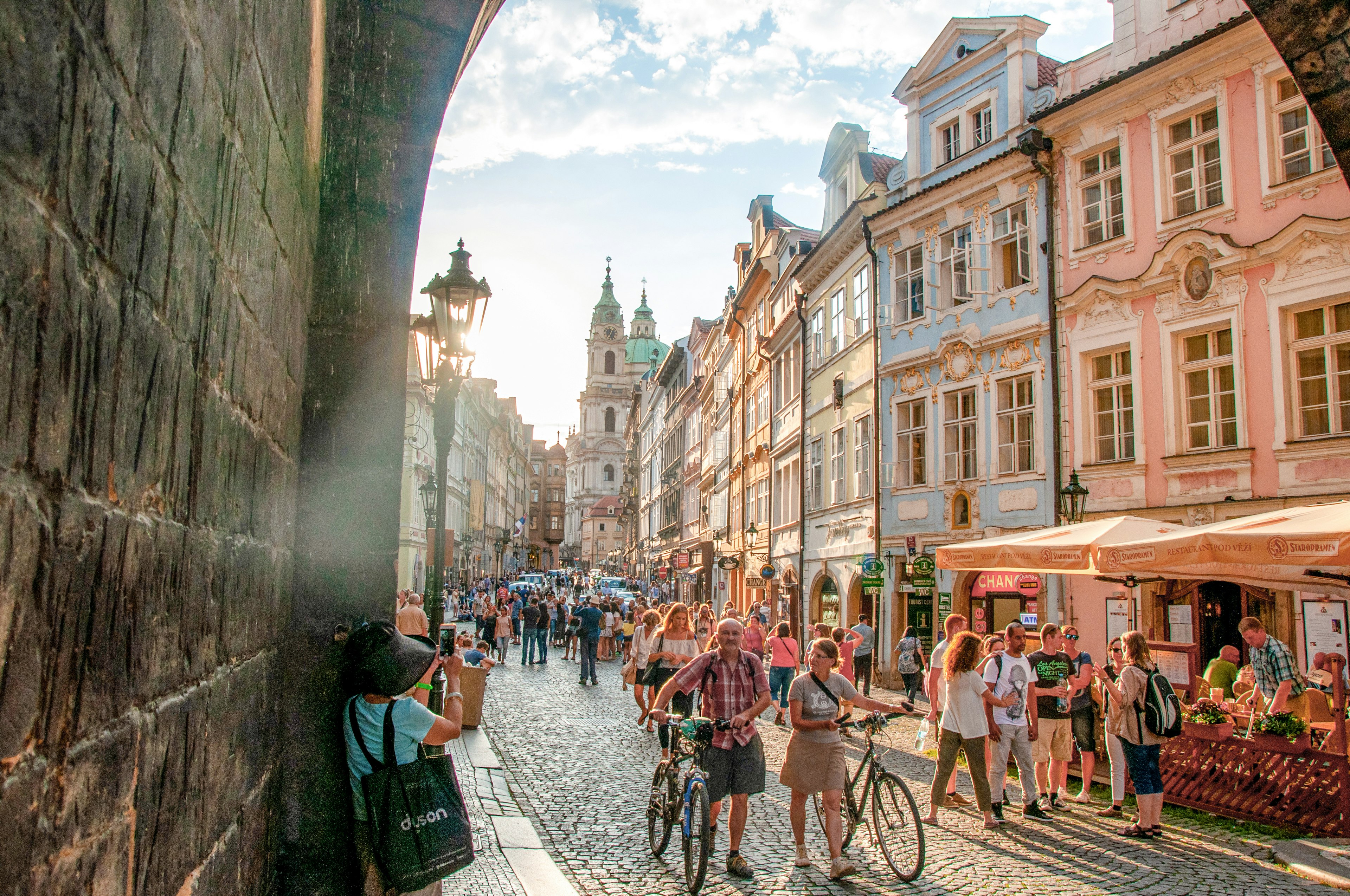 Many people are walking and pushing bicycles along a crowded cobblestone street in Prague at sunset in the summertime