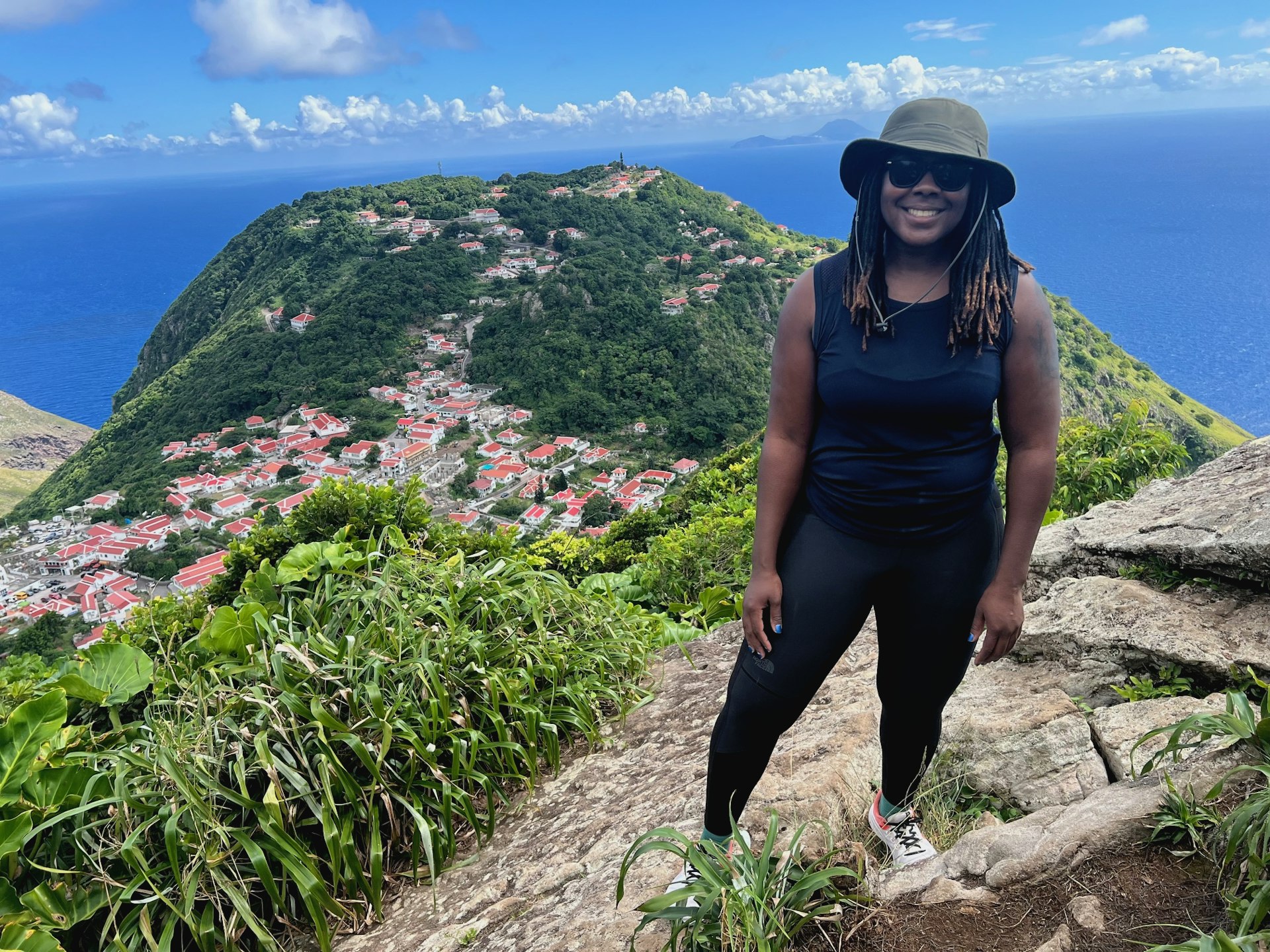 Lonely Planet Destination Editor Alicia Johnson faces the camera on the lookout point at Mas Cohones Hill on the Caribbean Island of Saba. In the background, there are the red rooftops of the Saban homes, a large hill and the expansive blue ocean. 