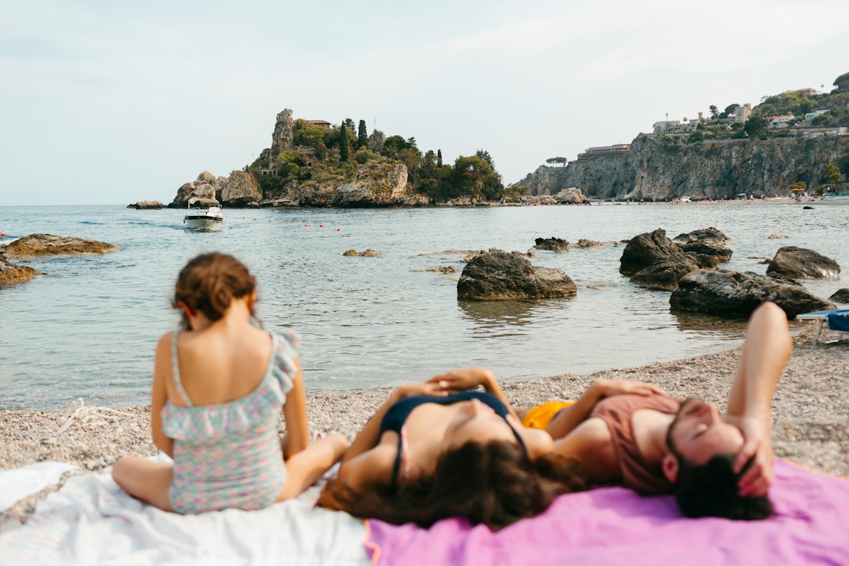 Young family on the shore of pebble beach lying down un sunlight, in front of Isola Bella island in SIcily, in the Mediterranean Sea