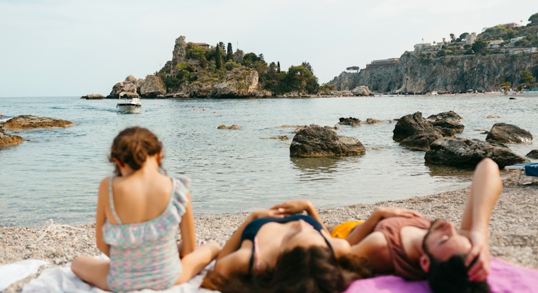 Young family on the shore of pebble beach lying down un sunlight, in front of Isola Bella island in SIcily, in the Mediterranean Sea