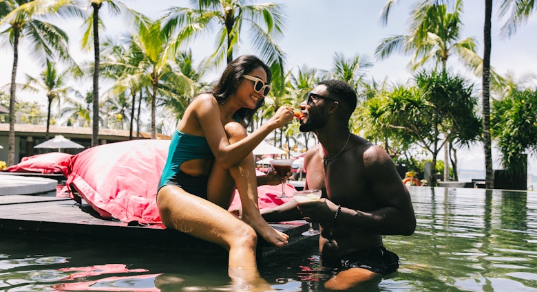 A mixed race couple having cocktails by the pool of a resort in the tropics. She is Asian, he is black. She is feeding him strawberries. Vacation concept.