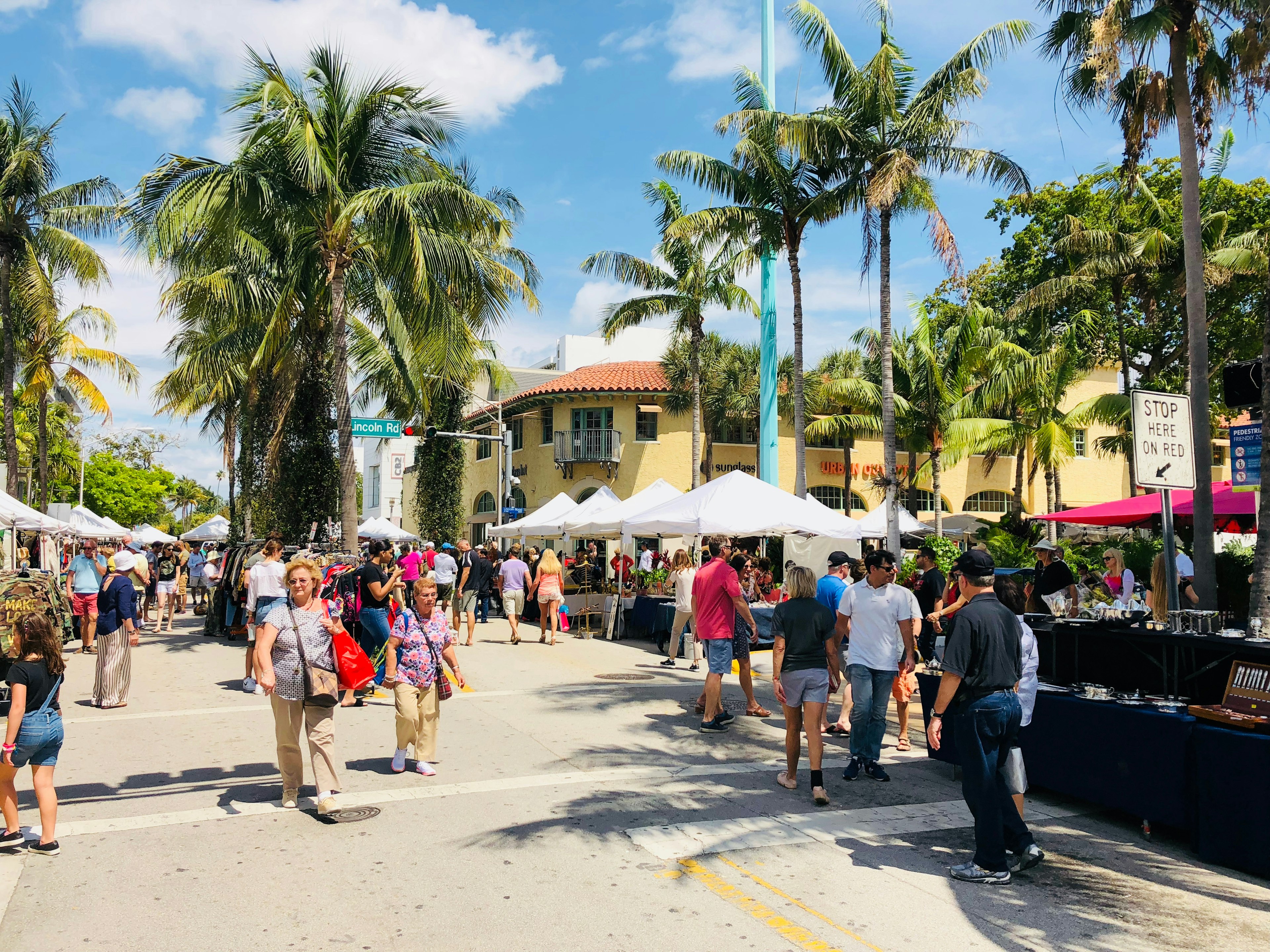 People wander among market stalls along a palm-lined street on a sunny day