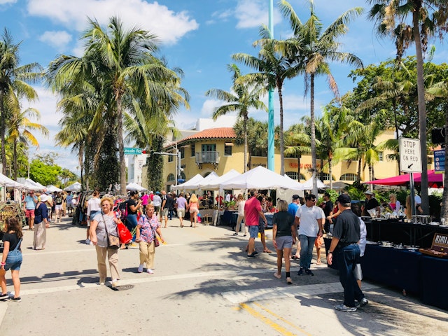 People wander among market stalls along a palm-lined street on a sunny day