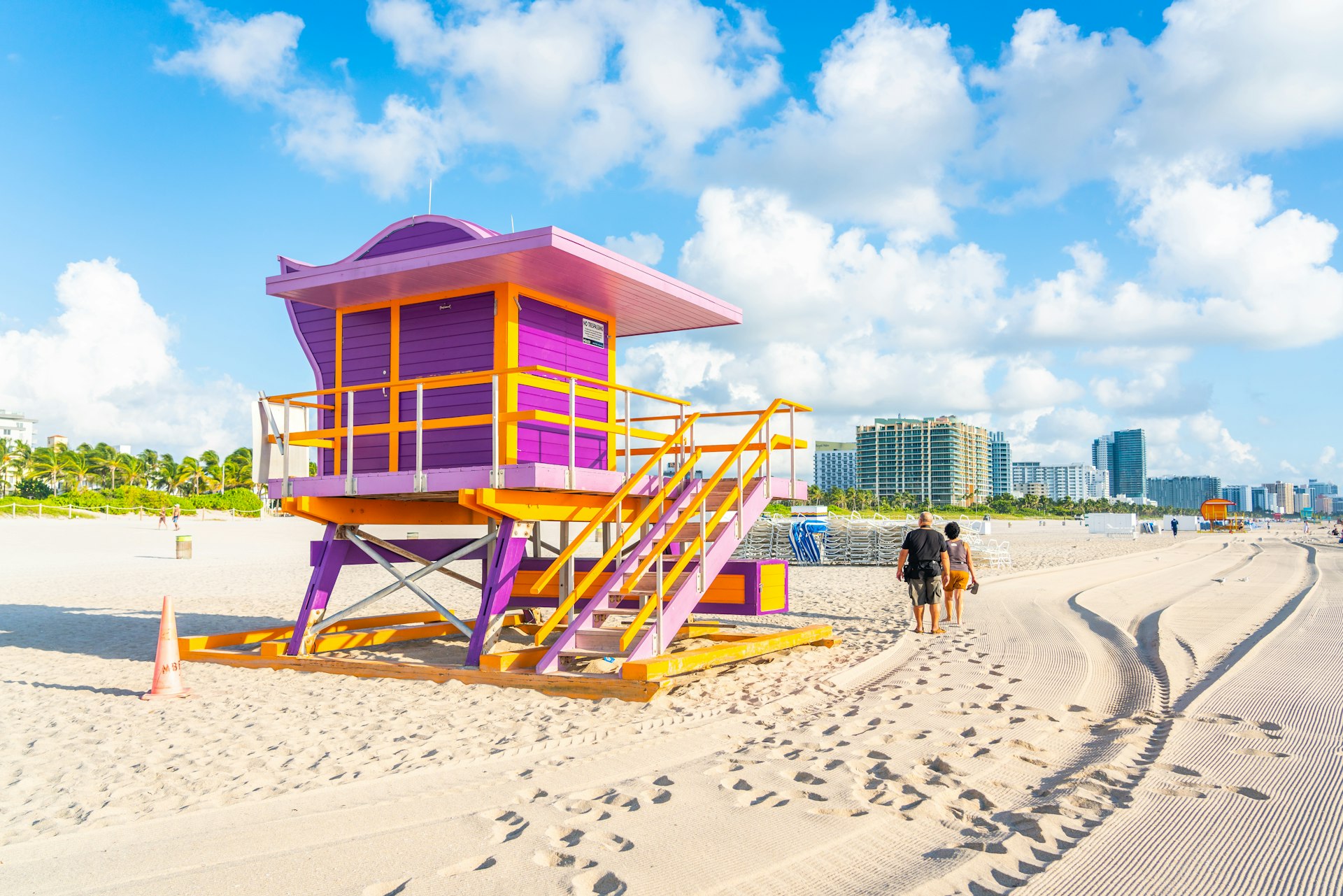 Two people walk by a pink and yellow art deco-style lifeguard hut on a sandy beach