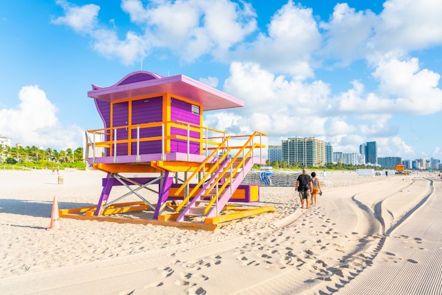 Two people walk by a pink and yellow art deco-style lifeguard hut on a sandy beach