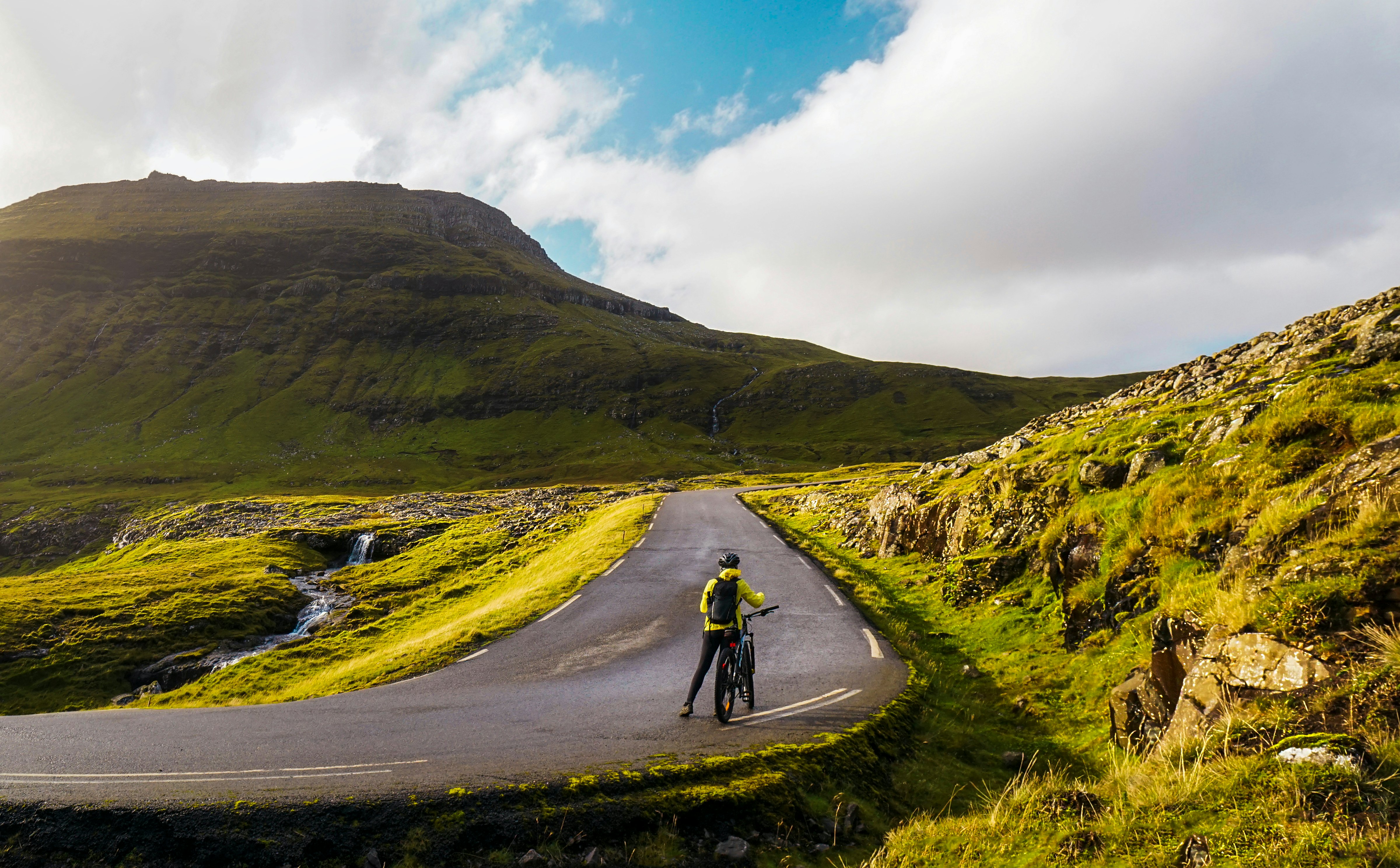 A cyclist stands next to their bike on a mountain road with beautiful green fields