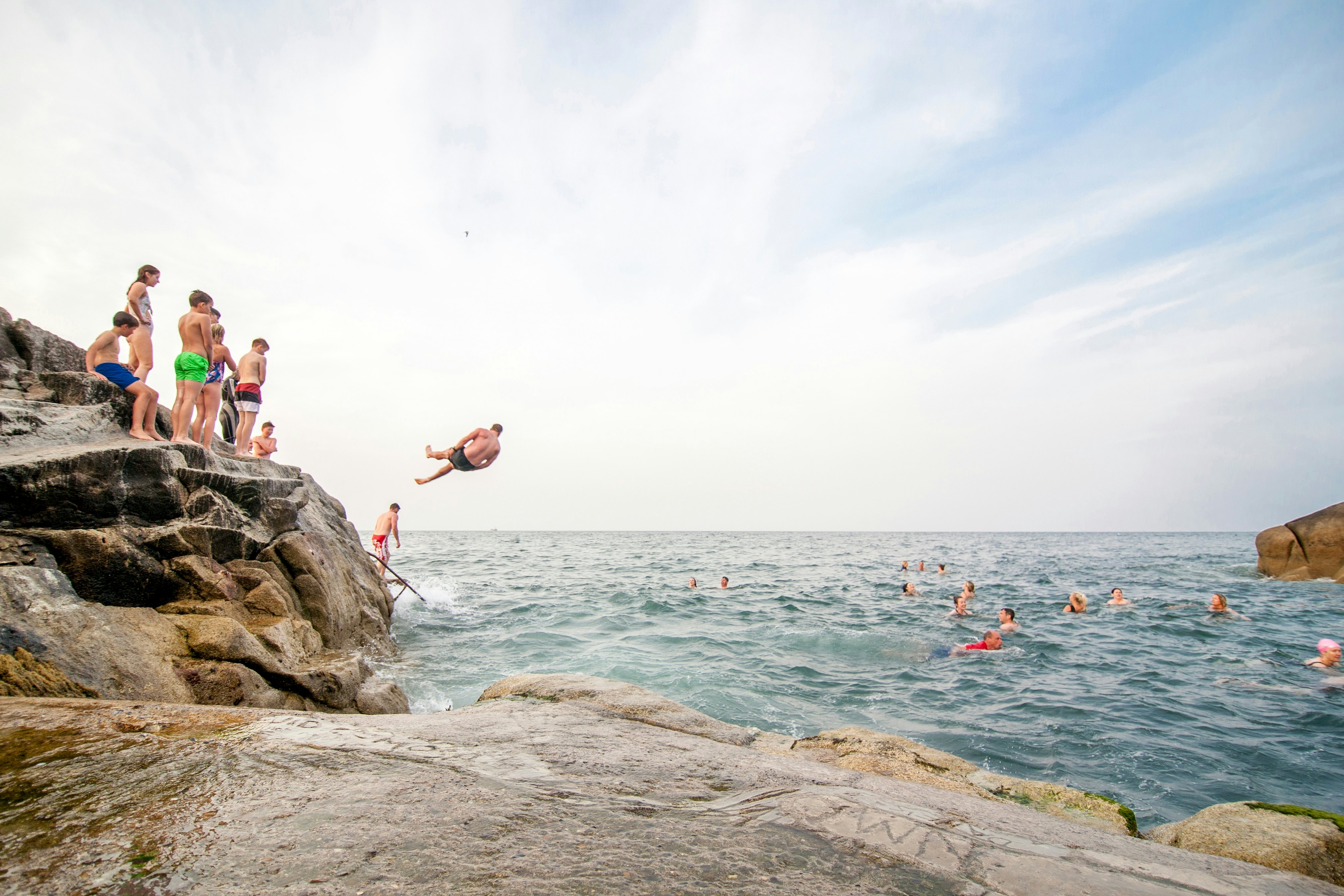 People line up to jump into the water from a small cliff. Other bathers are pictured swimming in a cove off the sea.