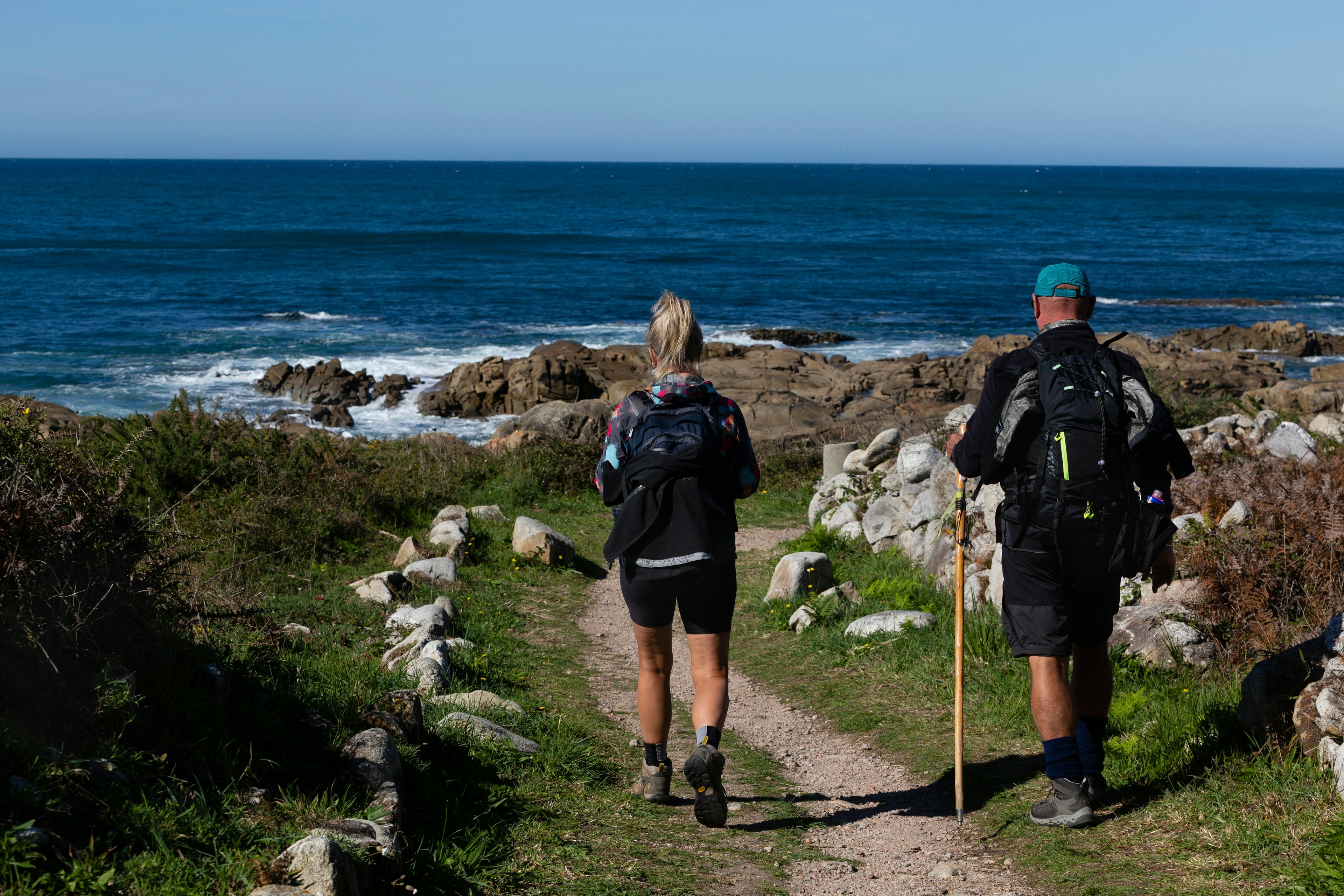 Two backpack-carrying hikers follow a rocky pathway beside the sea