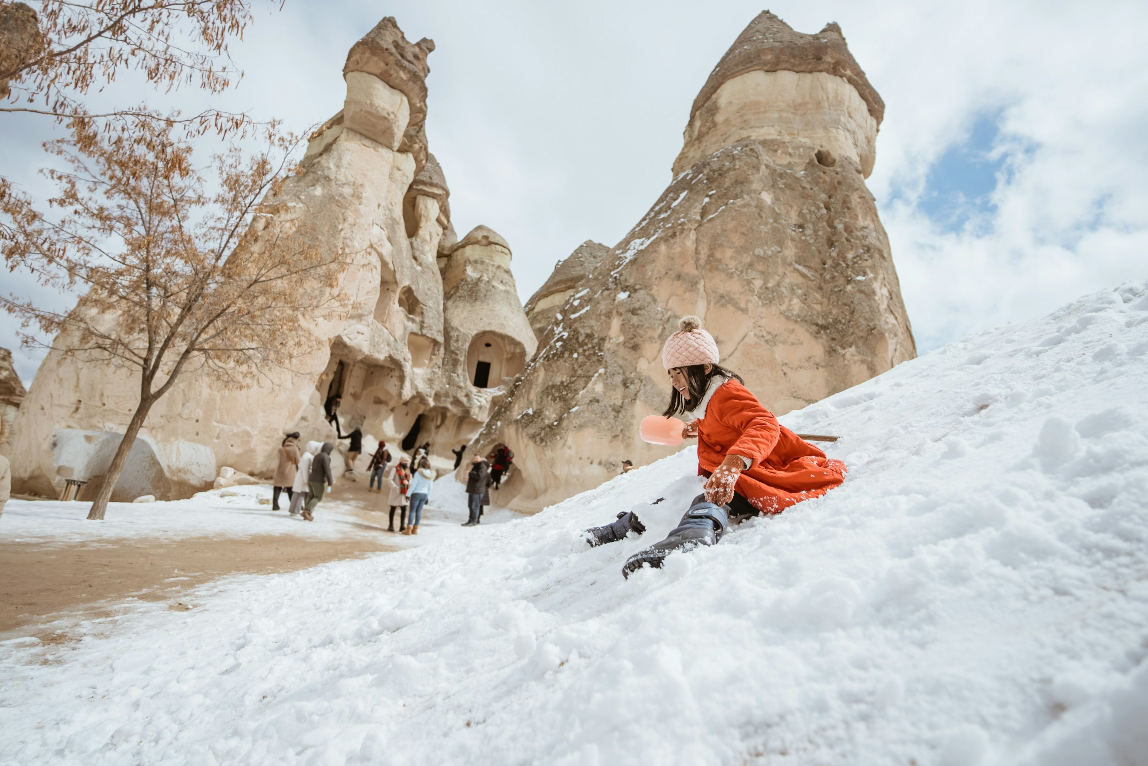 A little girl slides down the snow on a slope near some chimney-like rock formations.