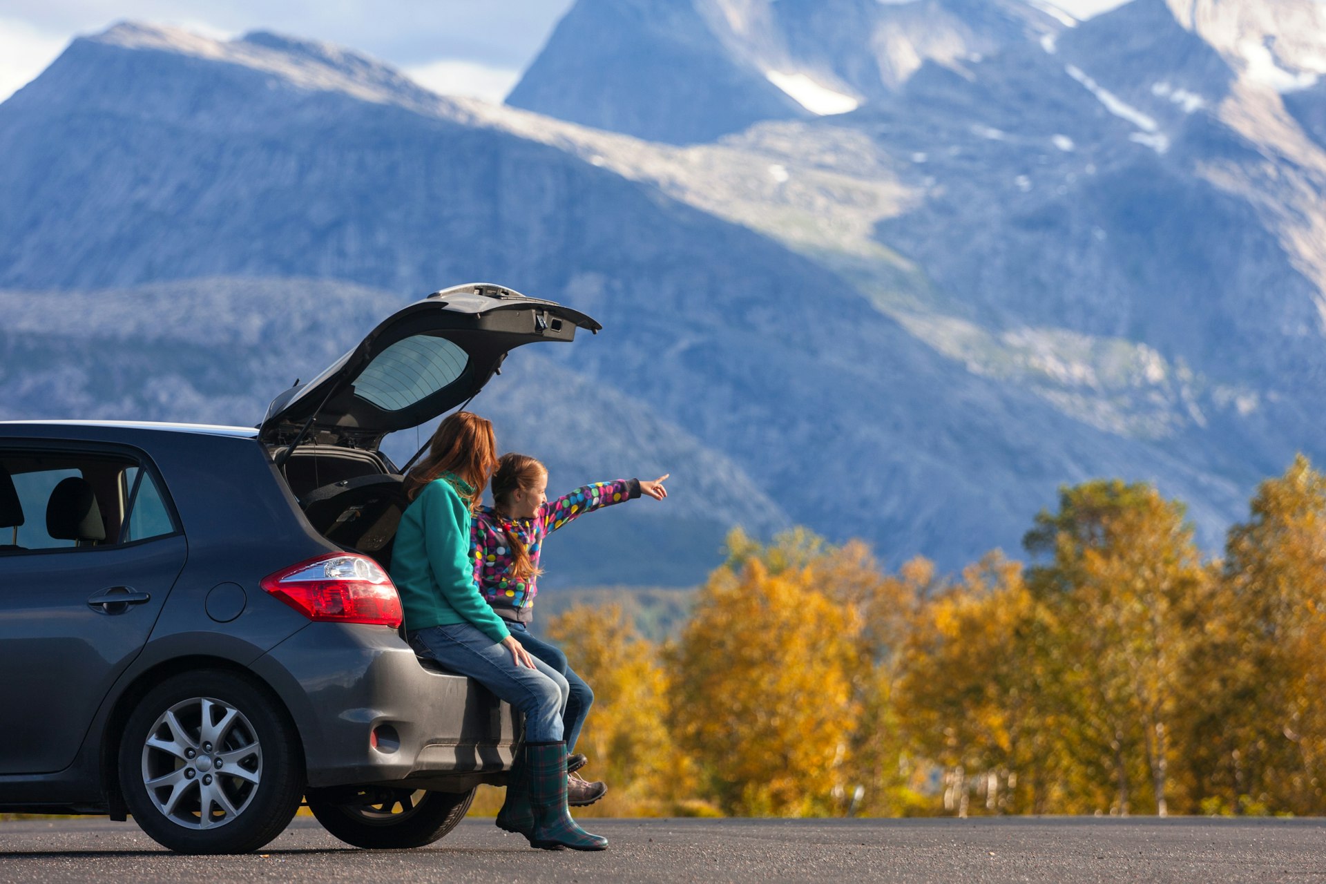 A mother and daughter sit on the tailgate of their SUV on the side of the road with mountain scenery in the distance, Norway