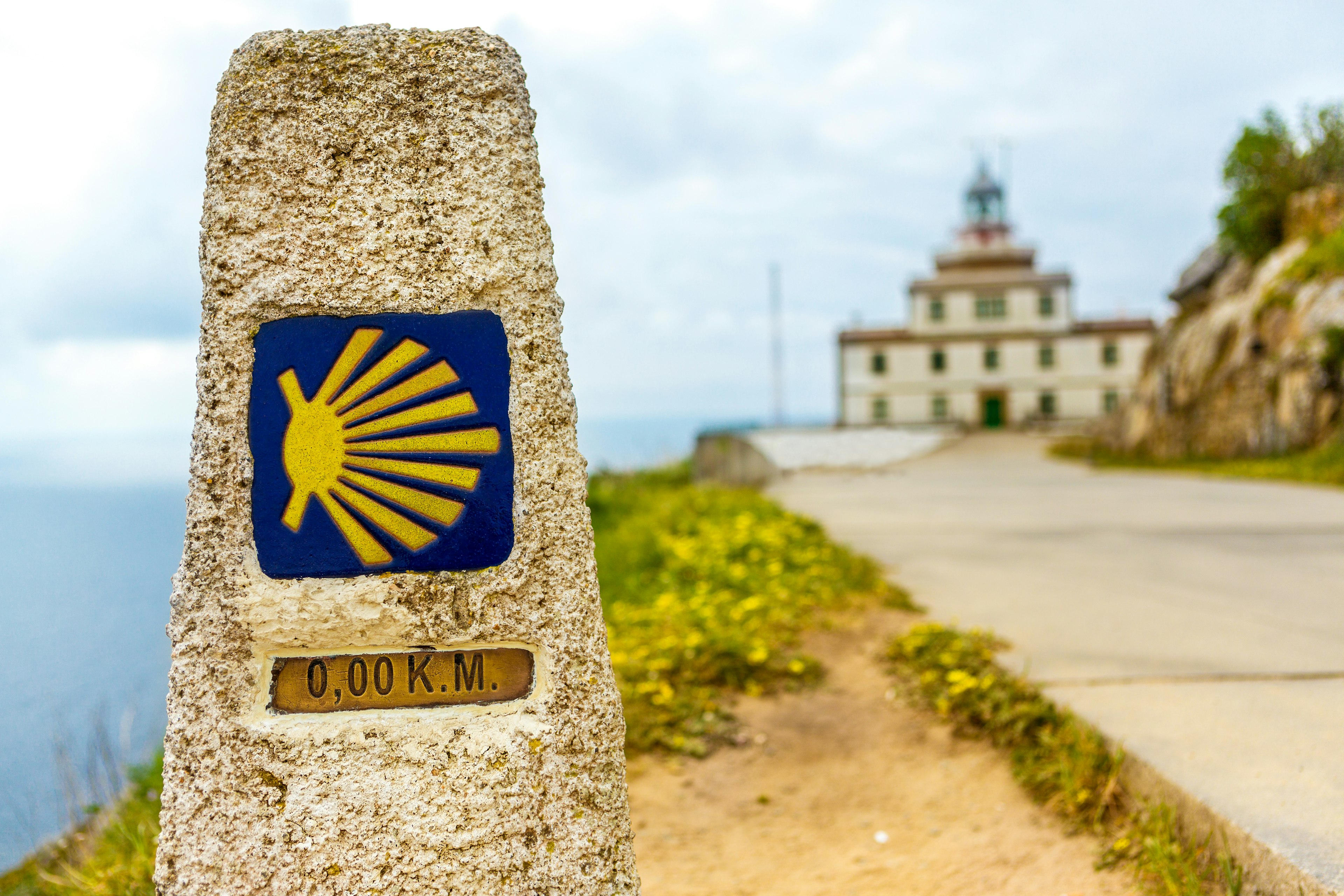 A 0.0km marker showing pilgrims the end of a route. There's a lighthouse in the distance.