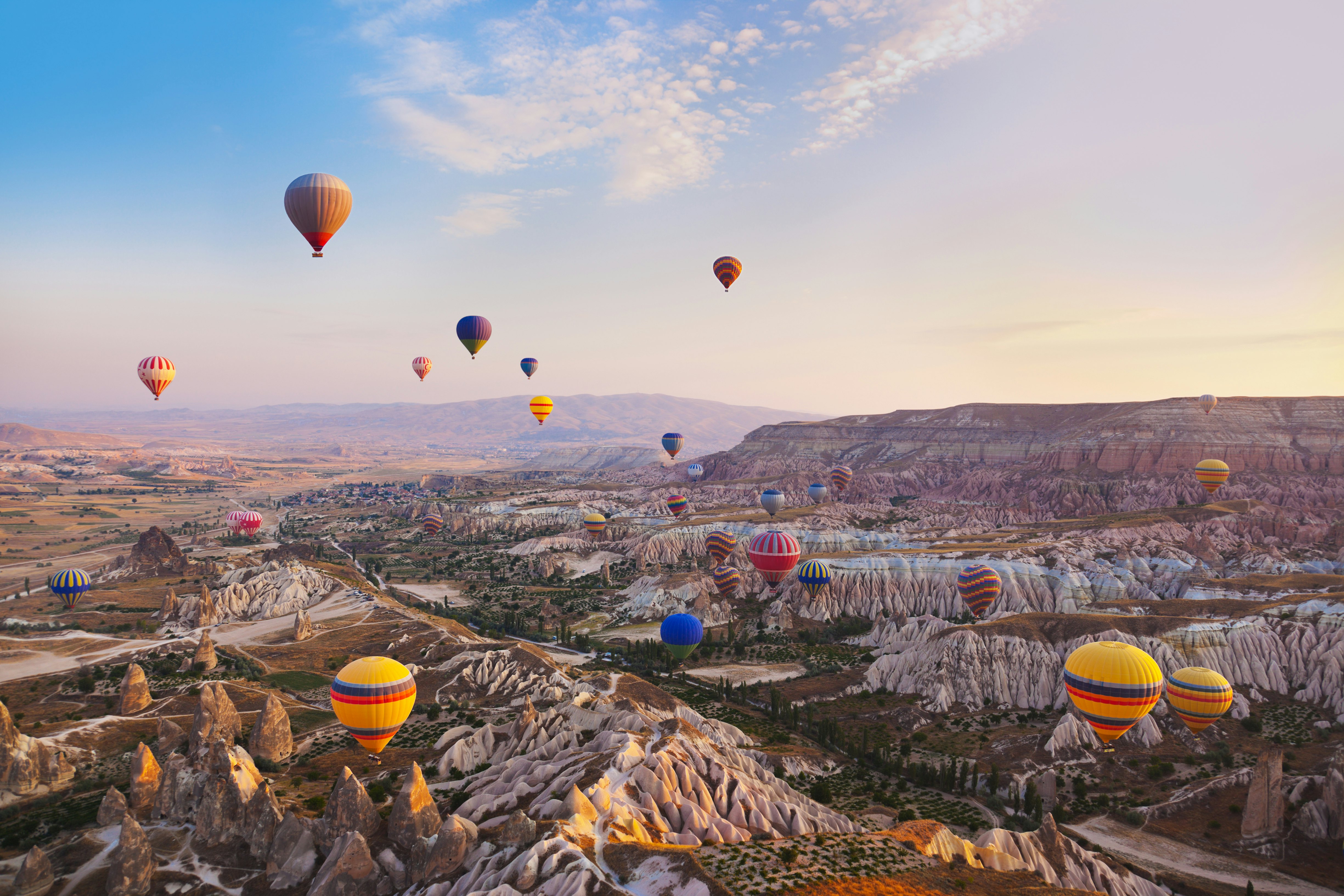 Multicolored hot-air balloons fly over a landscape with rock formations in Türkiye.