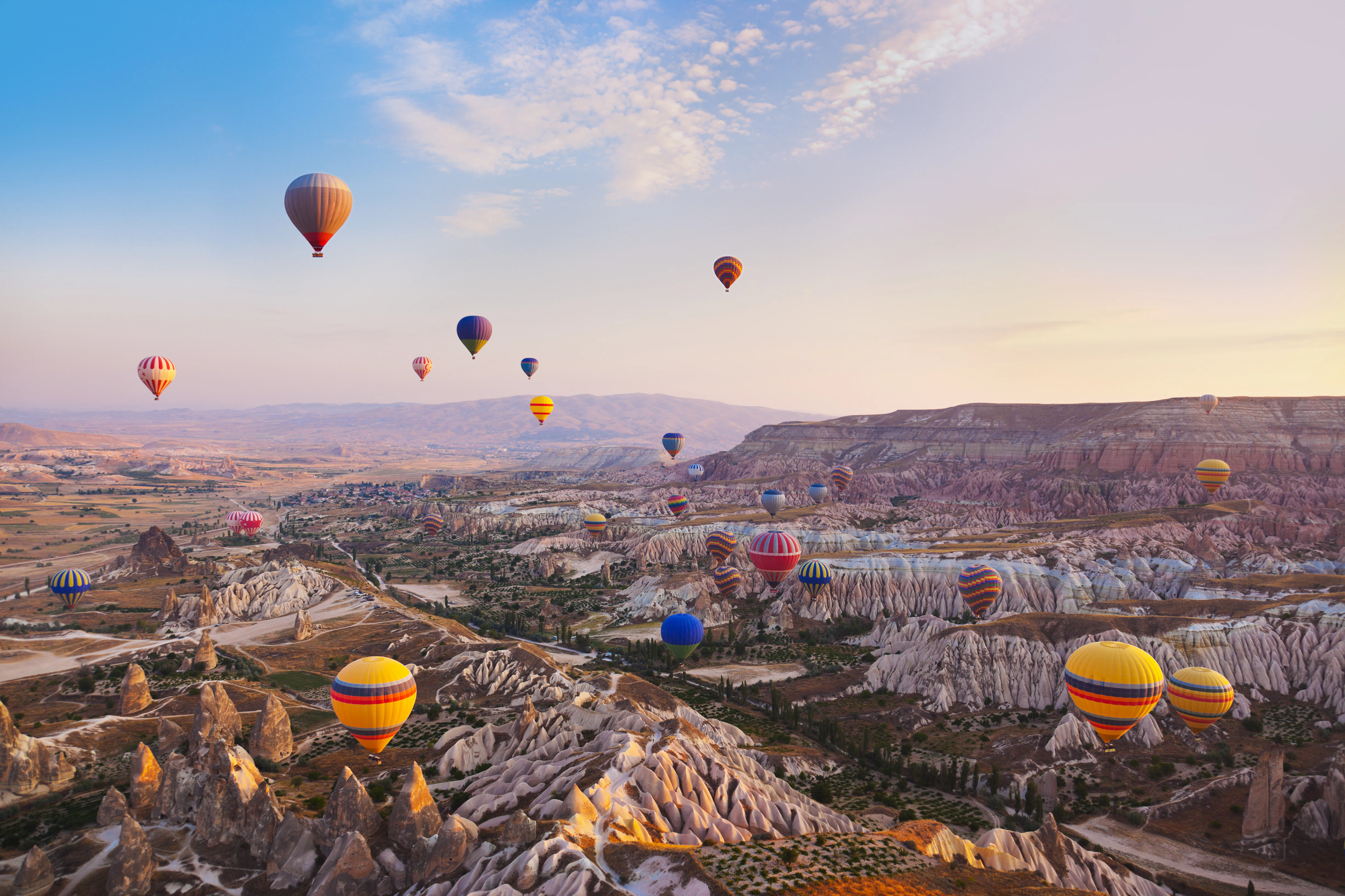 Multicolored hot-air balloons fly over a landscape with rock formations in Türkiye.