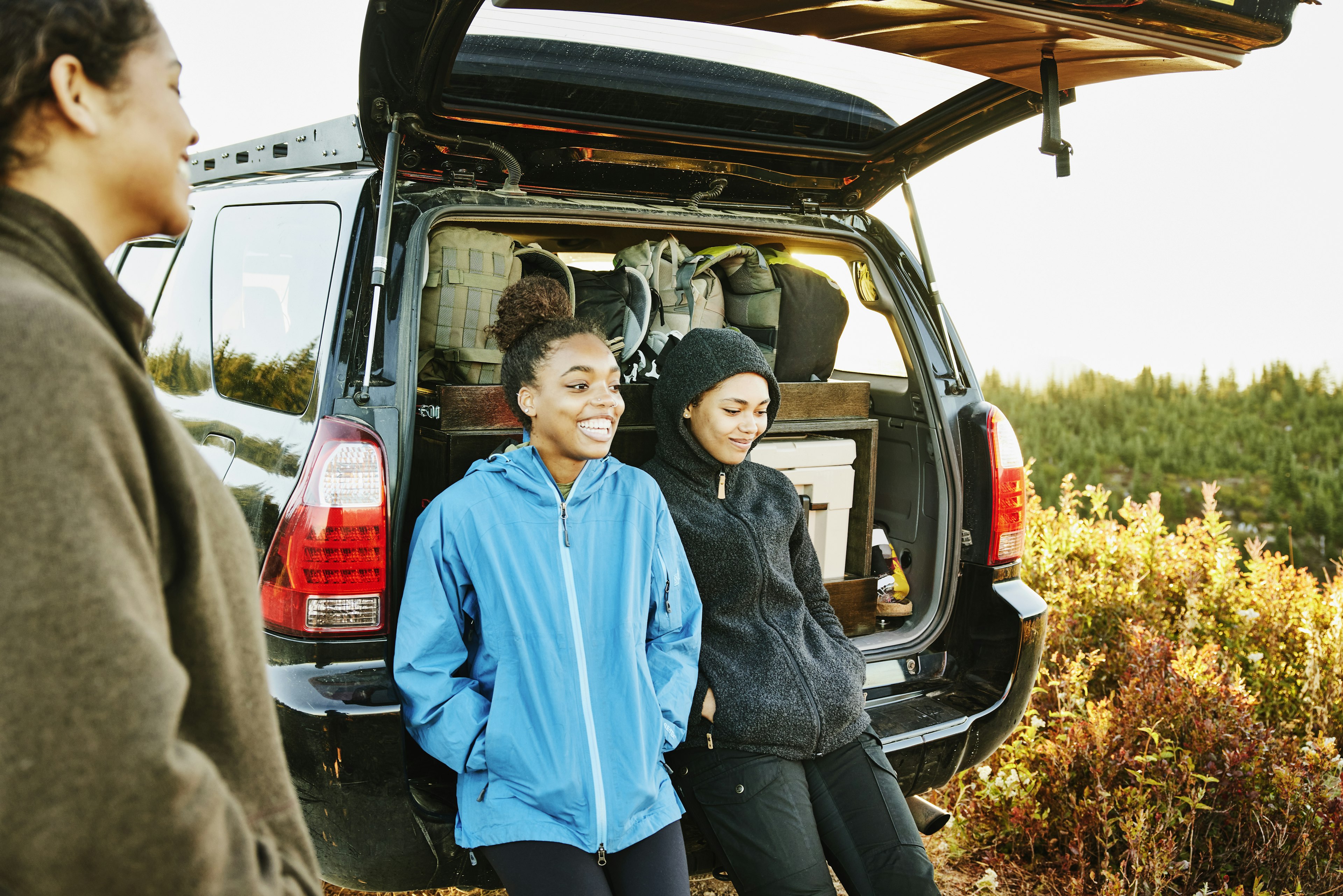 Smiling sisters hanging out at back of car after finishing backpacking trip