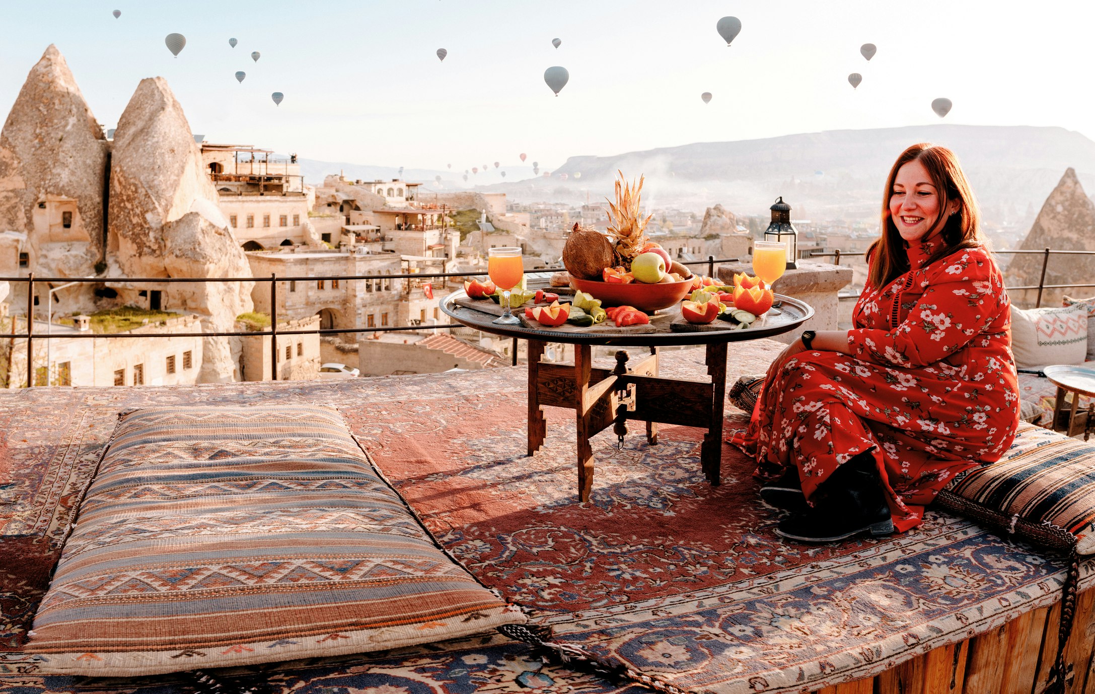 A woman on a hotel rooftop smiles at the beautiful scenery as hot-air-balloons soar in the sky above her
