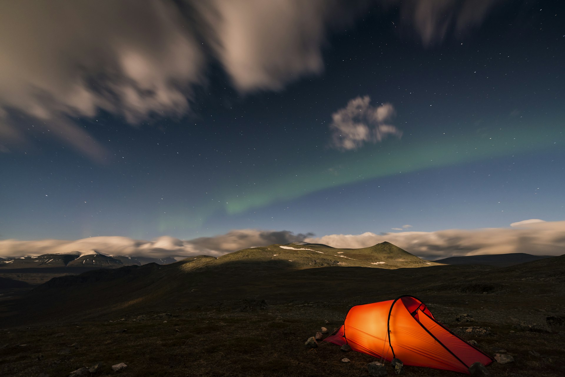 A tent lit up in orange under a night sky with a streak of green from the northern lights  