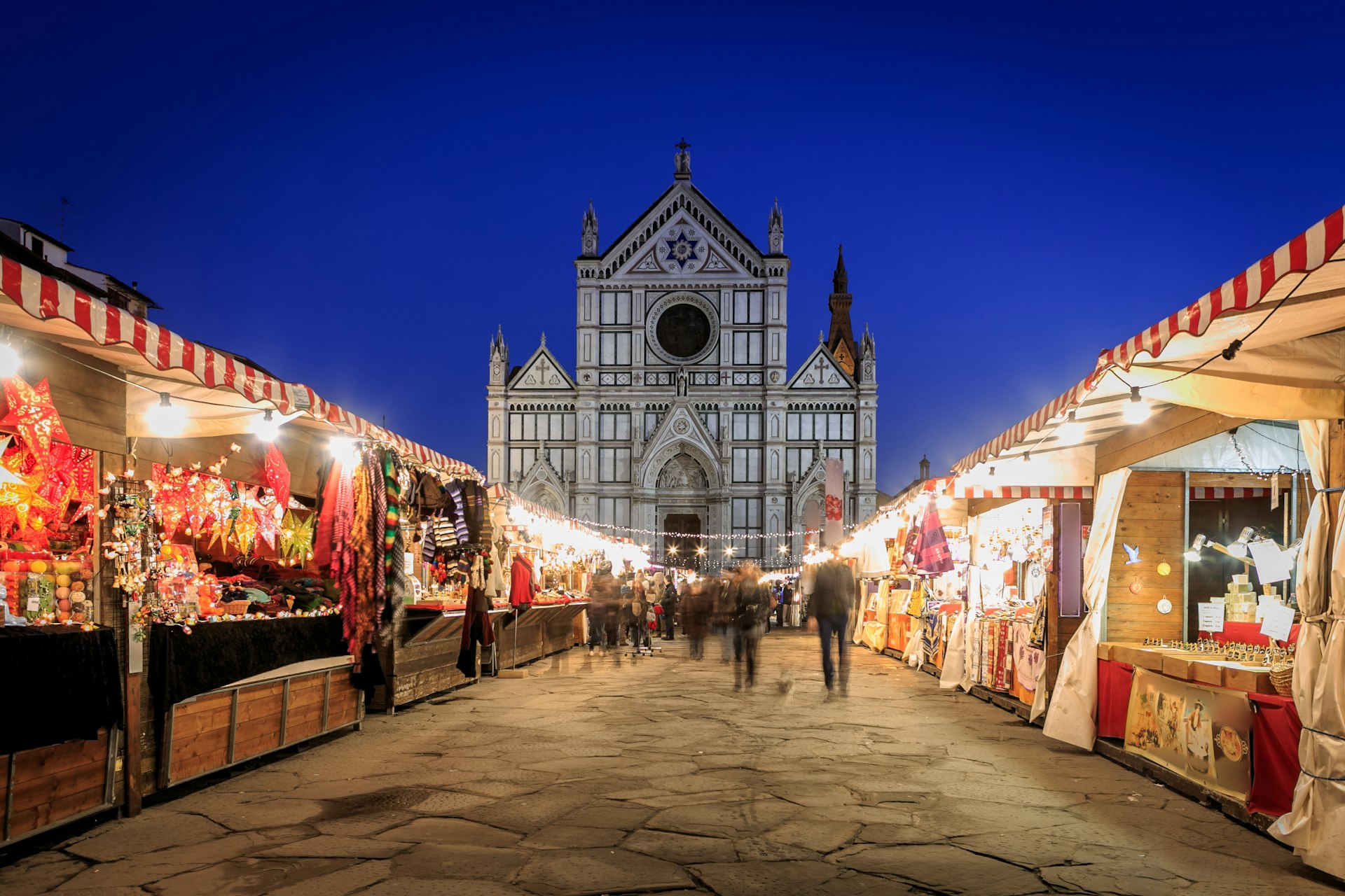 People shop at a row of winter market stalls lit up in front of a church
